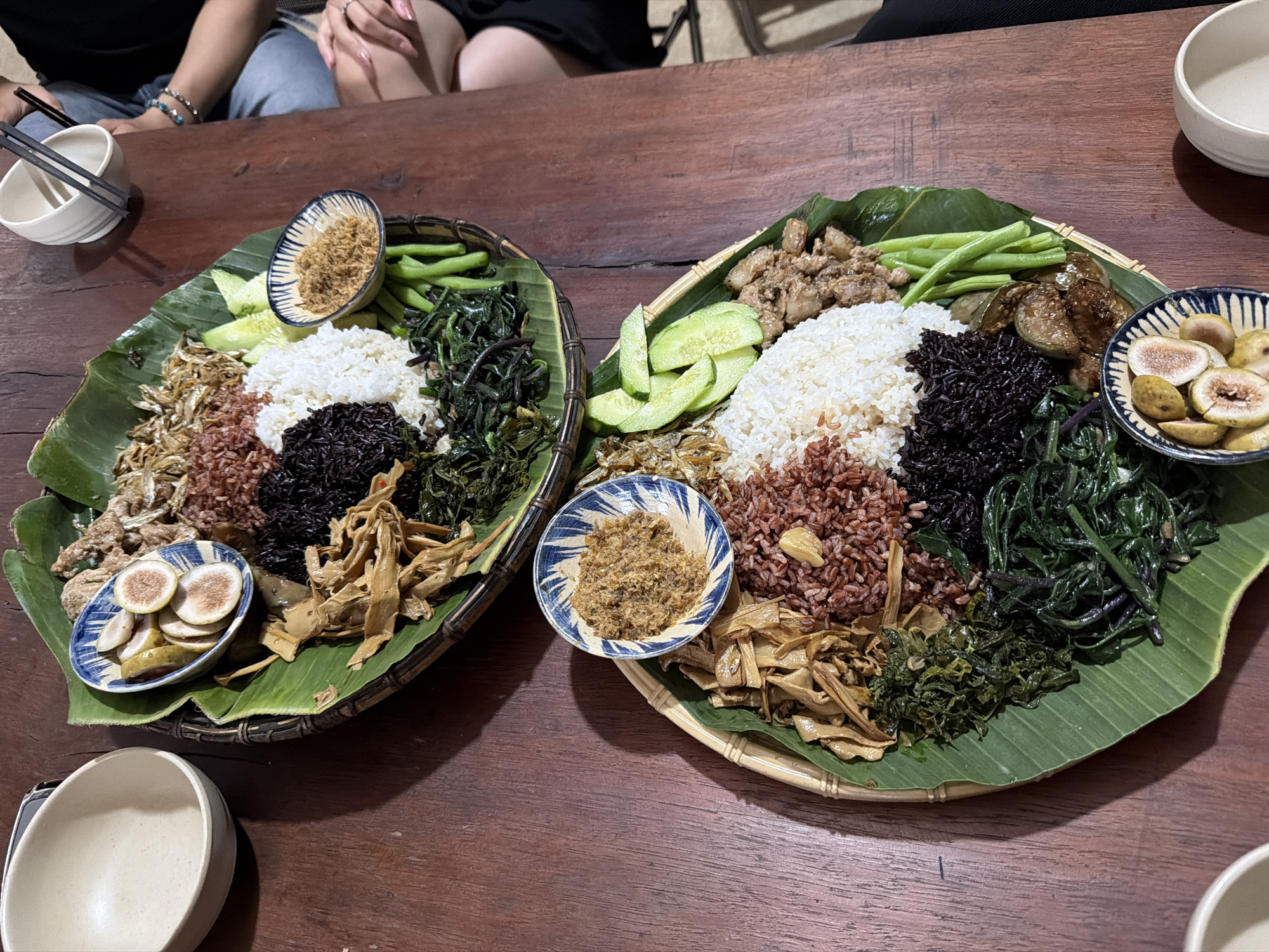 This overhead shot depicts a traditional Vietnamese meal served on two large, woven bamboo platters, each lined with fresh banana leaves. The platters are laden with a variety of colorful dishes, set on a dark wooden table, suggesting an evening dining experience.
Each platter features a central arrangement of three distinct types of rice: fluffy white rice, reddish-brown rice (likely brown or red rice), and dark purple or black rice. Surrounding the rice are numerous accompaniments. These include slices of fresh green cucumber, bright green beans, and generous portions of various cooked green leafy vegetables, some dark and leafy, others lighter. One platter also presents dark, segmented pieces resembling pickled or stewed eggplant.
Additional items on the platters include small dried fish or anchovies, chunks of what appears to be diced meat (possibly pork belly), and piles of light-brown, shredded dried bamboo shoots. Several small ceramic bowls with blue and white patterns contain condiments or side dishes. One bowl holds a fluffy, light brown substance resembling meat or fish floss (chà bông), while another contains pale, sliced items, possibly pickled unripe figs.
In the upper left background, parts of two individuals are visible, including their hands, arms adorned with bracelets, and legs, indicating they are seated at the table, about to partake in or are already enjoying the meal. A white ceramic bowl with chopsticks is placed next to the left food platter, and another empty white bowl is seen on the right. The communal presentation of food on large platters is typical of traditional dining in Măng Đen, Vietnam. No visible text is present in the image.