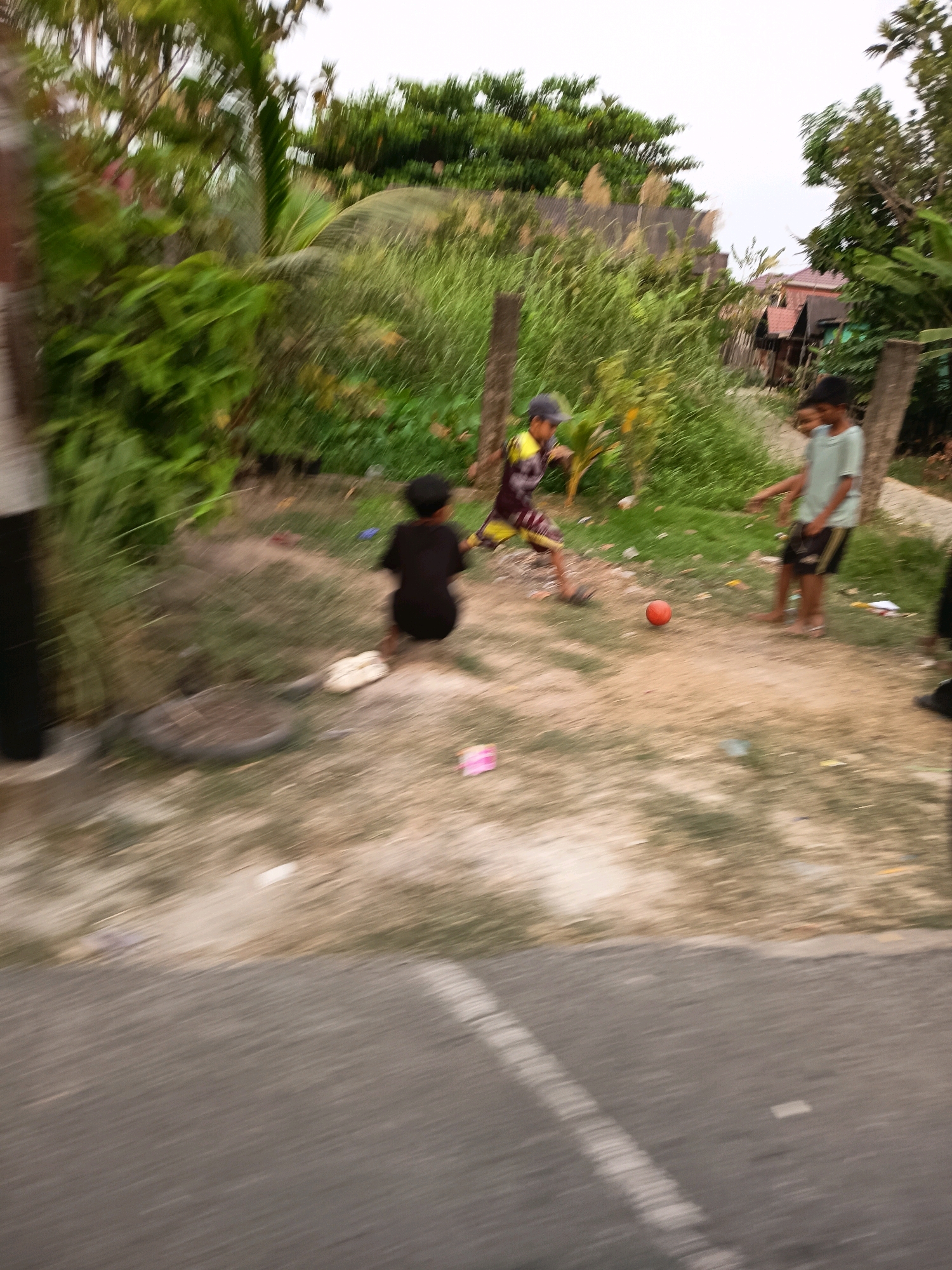 The image depicts an outdoor scene in Banjarmasin, Indonesia, likely a residential area with lush vegetation and some simple structures in the background. Three young boys appear to be playing with a red ball on a dirt and grass area adjacent to a road. One boy, wearing a dark t-shirt, is crouched down, seemingly about to kick the ball. Another boy, in a yellow and maroon patterned shirt and a cap, is in mid-action, also towards the ball. The third boy, in a light blue t-shirt, stands a short distance away, watching. The scene is captured with motion blur, suggesting the photograph was taken from a moving vehicle. The environment is characterized by overgrowth and a somewhat untidy appearance, typical of many urban fringe areas in the region.