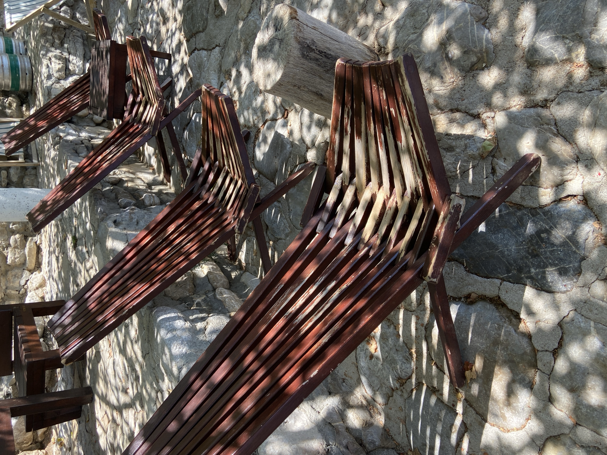 In Prijevor, Montenegro, a cluster of weathered, dark brown wooden lounge chairs is arranged on a cobblestone patio. Sunlight casts dappled shadows across the scene, highlighting the peeling paint on the chairs and the textured stones underfoot. The chairs, designed with multiple slats, are reclined, suggesting a space intended for relaxation. In the background, a stone wall rises, and further beyond, several metallic kegs are visible, hinting at a nearby establishment, possibly a bar or restaurant. A rough-hewn log sits to the right of the chairs, serving as a rustic element in the outdoor seating area. The overall impression is one of a casual, sun-drenched outdoor space.