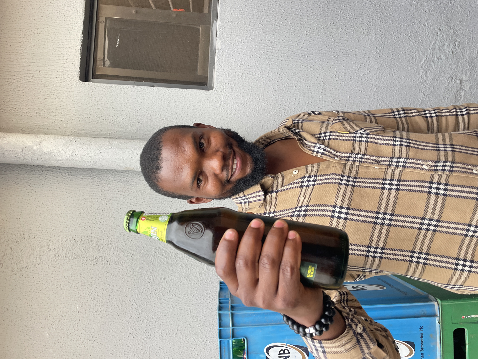 A man with a dark complexion, wearing a brown and black checkered button-down shirt and a black beaded bracelet, is holding a bottle of Harp Lager beer. He is smiling and looking towards the camera. He appears to be standing next to a stack of blue and green plastic bottle crates. The background consists of a light gray wall, and there is a window visible in the upper portion of the image. Although asked, I cannot provide the location in the city of Jos, Nigeria, as I do not have the information needed to fulfill that specific request.