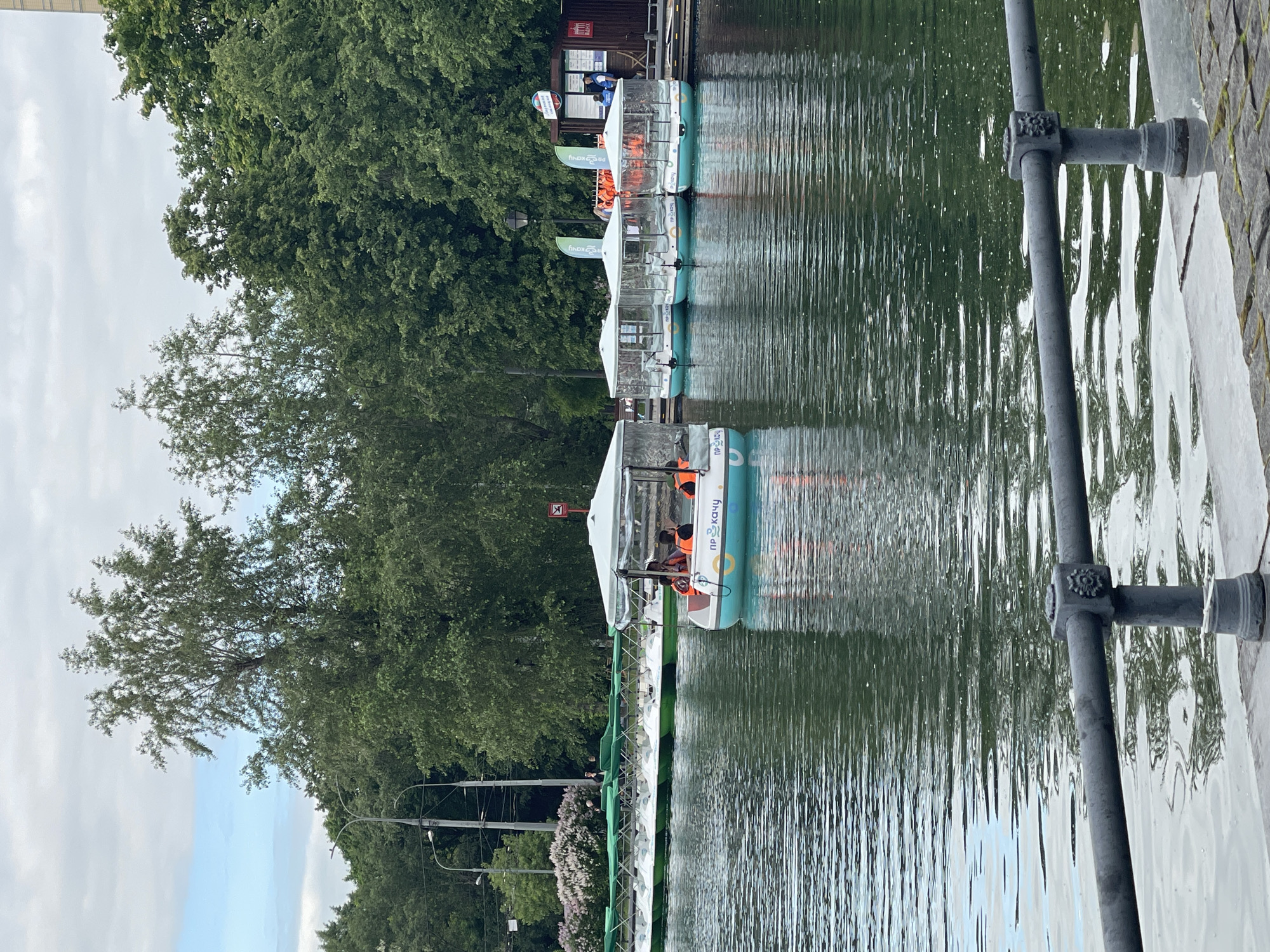 The image captures a serene daytime scene at a boat rental dock on a body of water, likely a lake or river, under a partly cloudy sky. Several pedal boats or paddle boats are moored in a line along a wooden dock.
In the foreground, the calm water shows gentle ripples, reflecting the sky and surrounding greenery. To the right, a dark gray metal railing with decorative elements separates the water from a paved, possibly stone, walkway or dock edge.
The main subjects are the white and teal-colored pedal boats. At least five boats are visible, each equipped with a clear canopy. Some of the boats have individuals or small groups inside, wearing bright orange life jackets, suggesting a recreational activity. On the side of the boats, the Cyrillic text "прокачу" (prokachu), which translates to "I'll ride" or "I'll give a ride," is visible, indicating a rental service. A red and white sign with a crossed-out symbol is also attached to one of the boats.
Behind the boats, lush green trees line the bank, contributing to the natural setting. In the background, nestled among the trees, there's a small wooden structure, possibly a kiosk or information booth, with "БАНК" (BANK) and other indistinct Cyrillic text visible on its facade. Further to the left, electrical poles and lines are discernible behind more trees, and a patch of flowering bushes, possibly lilac, adds a touch of color. The overall atmosphere is peaceful and leisurely, characteristic of a park or recreational area.