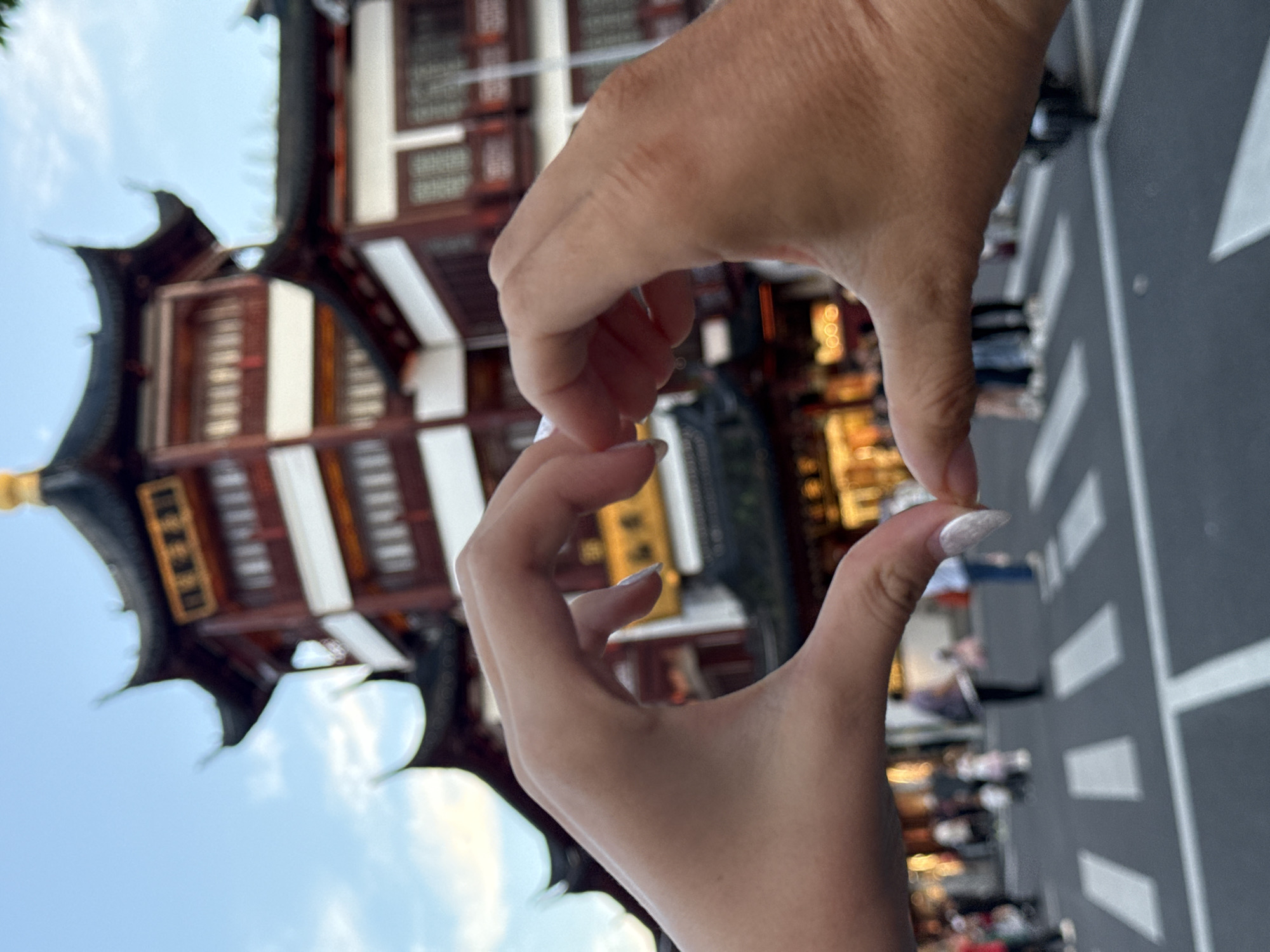 This is a close-up, eye-level, outdoor photograph taken in Shanghai, China, likely at dusk. In the foreground, two hands are forming a heart shape. The left hand, with manicured fingernails, is on the left side of the frame, and the right hand, with more pronounced skin texture, is on the right. Both hands are oriented towards the viewer, with thumbs and index fingers meeting to create the top curves of the heart, and the remaining fingers forming the bottom point.
In the midground, a pedestrian street is visible, marked by a crosswalk with white stripes. Several people are walking in the background, appearing as blurred figures. The architecture in the background is distinctly traditional Chinese, with multiple tiered roofs featuring dark, curved eaves and red and white painted facades. A golden finial crowns the tallest structure. The sky above is a pale blue with scattered white clouds. The overall impression is a tourist capturing a moment of affection with a scenic backdrop of a historic Chinese building complex.