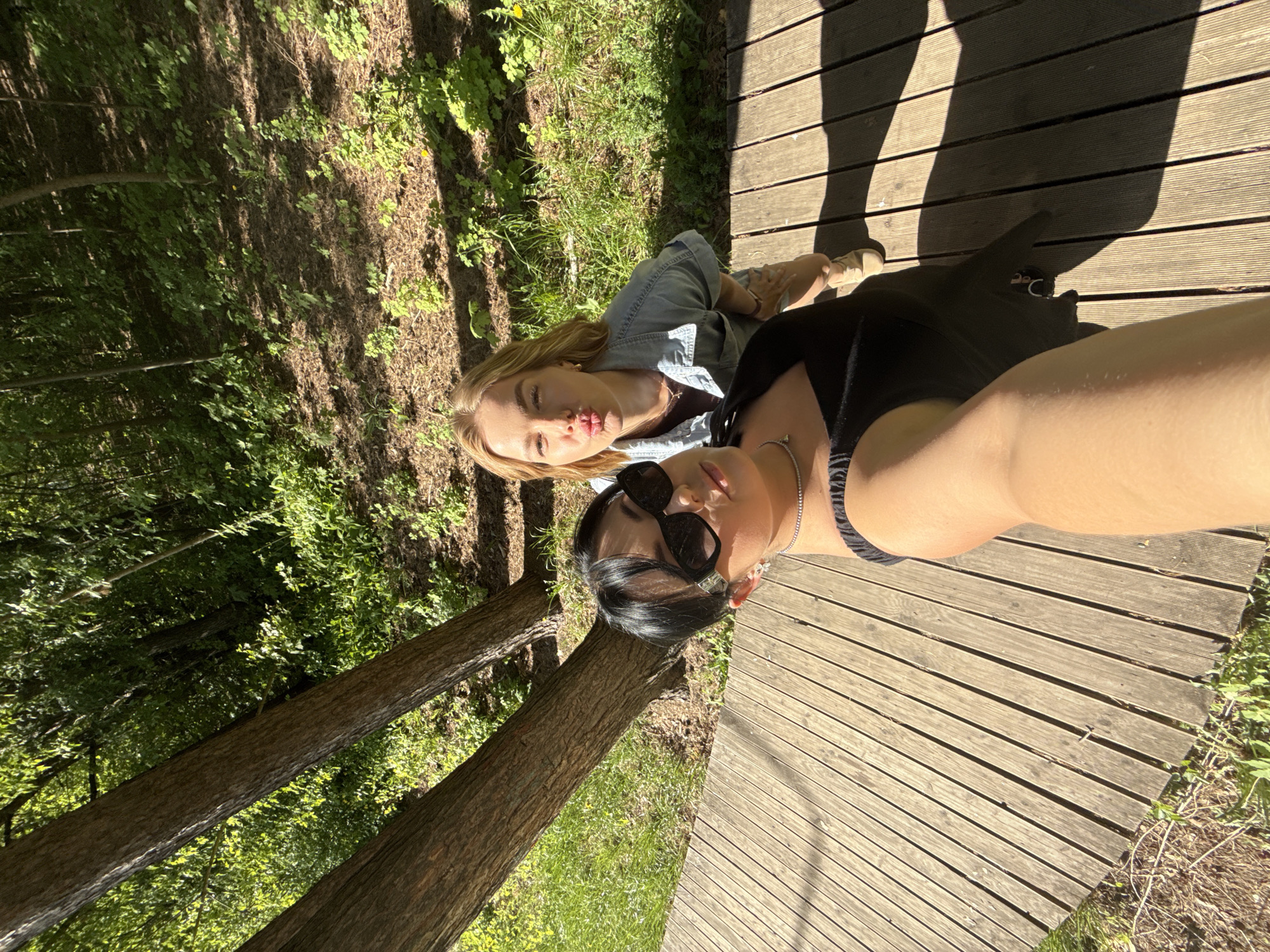 Two women are posing for a selfie on a wooden walkway in a park or wooded area. The woman on the left has blonde hair and is wearing a denim shirt. The woman on the right has black hair and is wearing sunglasses and a black top. Trees and foliage surround the walkway. The location is likely in Chaikovsky, Russia, based on the prompt.