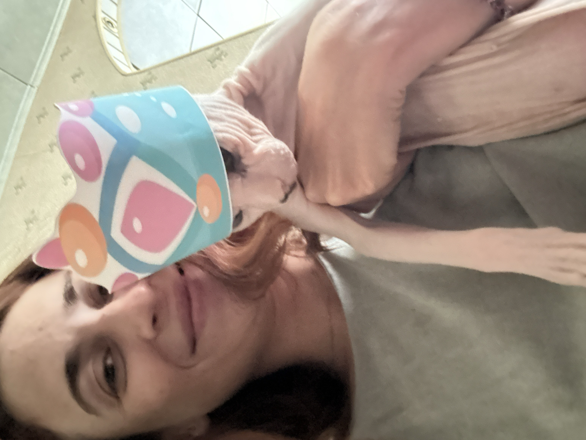 The image depicts a close-up selfie of a person holding a hairless cat. The person, with reddish-brown hair, is smiling and wearing a grey t-shirt. The cat, also hairless, is wearing a light blue party hat with colorful circles and shapes. The background appears to be an indoor setting, possibly a home, with patterned wallpaper and a hint of a mirror or window. There is no identifiable information in the image to suggest a location in Togliatti, Russia.