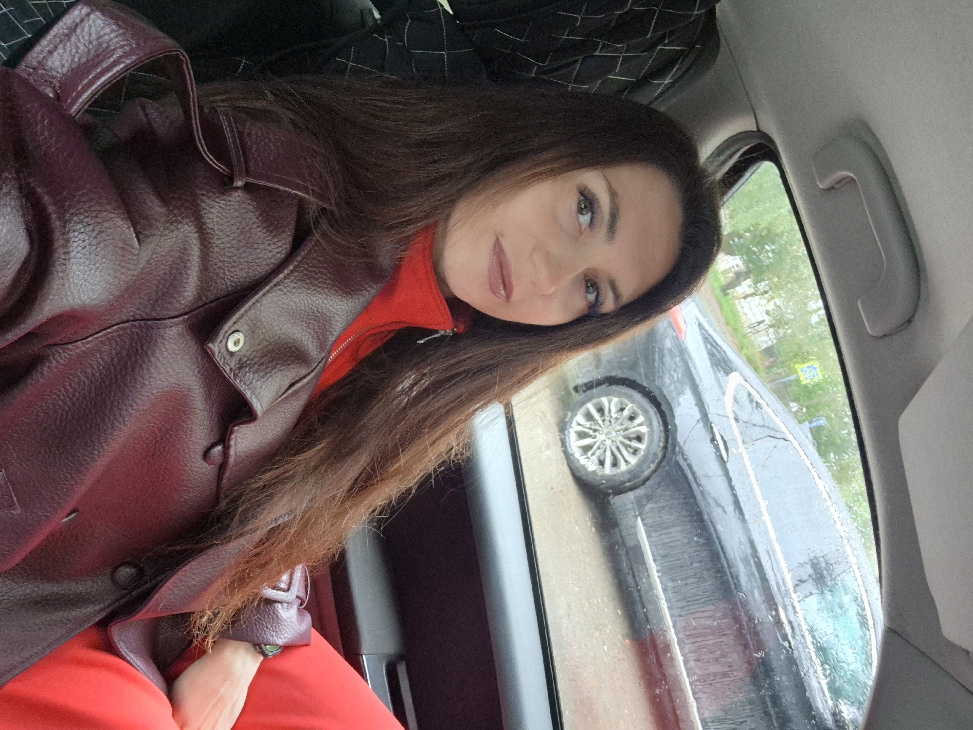 The image depicts a woman sitting in the back seat of a car. She has long, dark brown hair and is wearing a red top and a burgundy leather jacket. She is looking towards the camera with a neutral expression. Outside the car window, another black car is visible, and it appears to be raining, as evidenced by the water droplets on the car window. The background suggests an urban or suburban setting with some trees and buildings visible. The scene is likely captured in Chaikovsky, Russia, given the provided context.