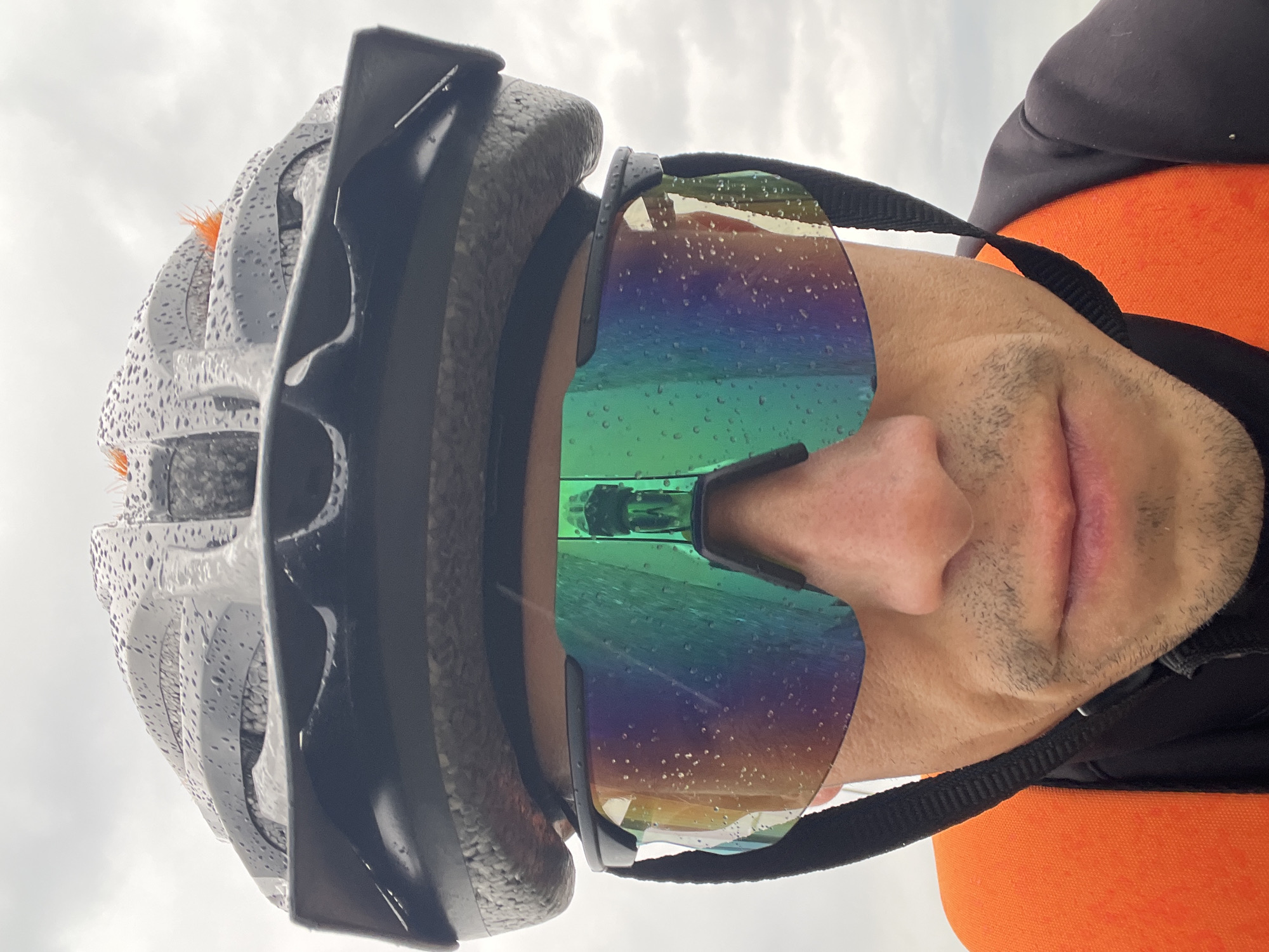 This close-up, low-angle image, taken in Chaikovsky, Russia, features a person wearing cycling gear in what appears to be rainy weather. The subject's head is tilted upwards, showing the lower half of their face, including the nose, mouth, and chin with light stubble.
They are wearing a grey or white cycling helmet, which is visibly covered in numerous water droplets, suggesting recent rain. Below the helmet and partially covered by a black outer layer, an orange garment is visible. Dominating the center of the image are large, wraparound cycling sunglasses with iridescent, rainbow-tinted lenses, also heavily speckled with water droplets. A black strap, likely part of the helmet or sunglasses, runs across the side of the face.
The background is a uniform, overcast grey sky, reinforcing the impression of a cloudy and wet outdoor environment. The image captures a moment, possibly during or after a bike ride, with the subject facing the inclement weather. No specific activity beyond being equipped for cycling in the rain is depicted, and there is no visible text.
