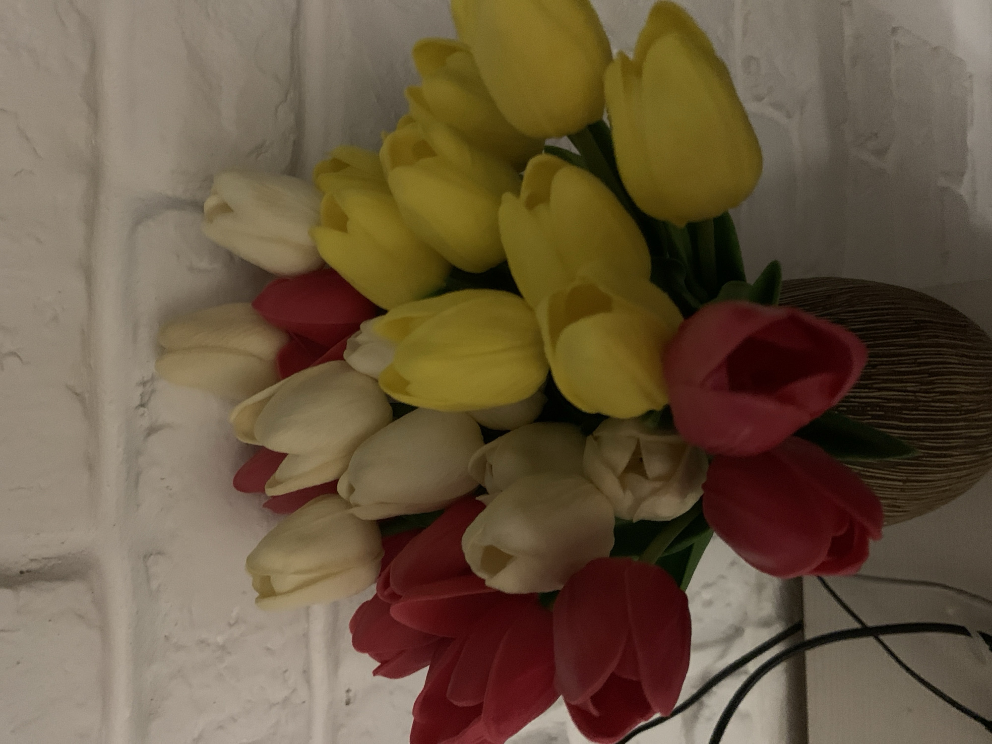 A bouquet of artificial tulips in red, yellow, and cream colors is displayed in a textured brown vase against a white brick wall. The tulips are arranged in a dense cluster, with some buds and fully opened blooms visible. The lighting in the scene is dim, casting soft shadows and highlighting the textures of the flowers and the brick wall. The location is not specified in the image.
