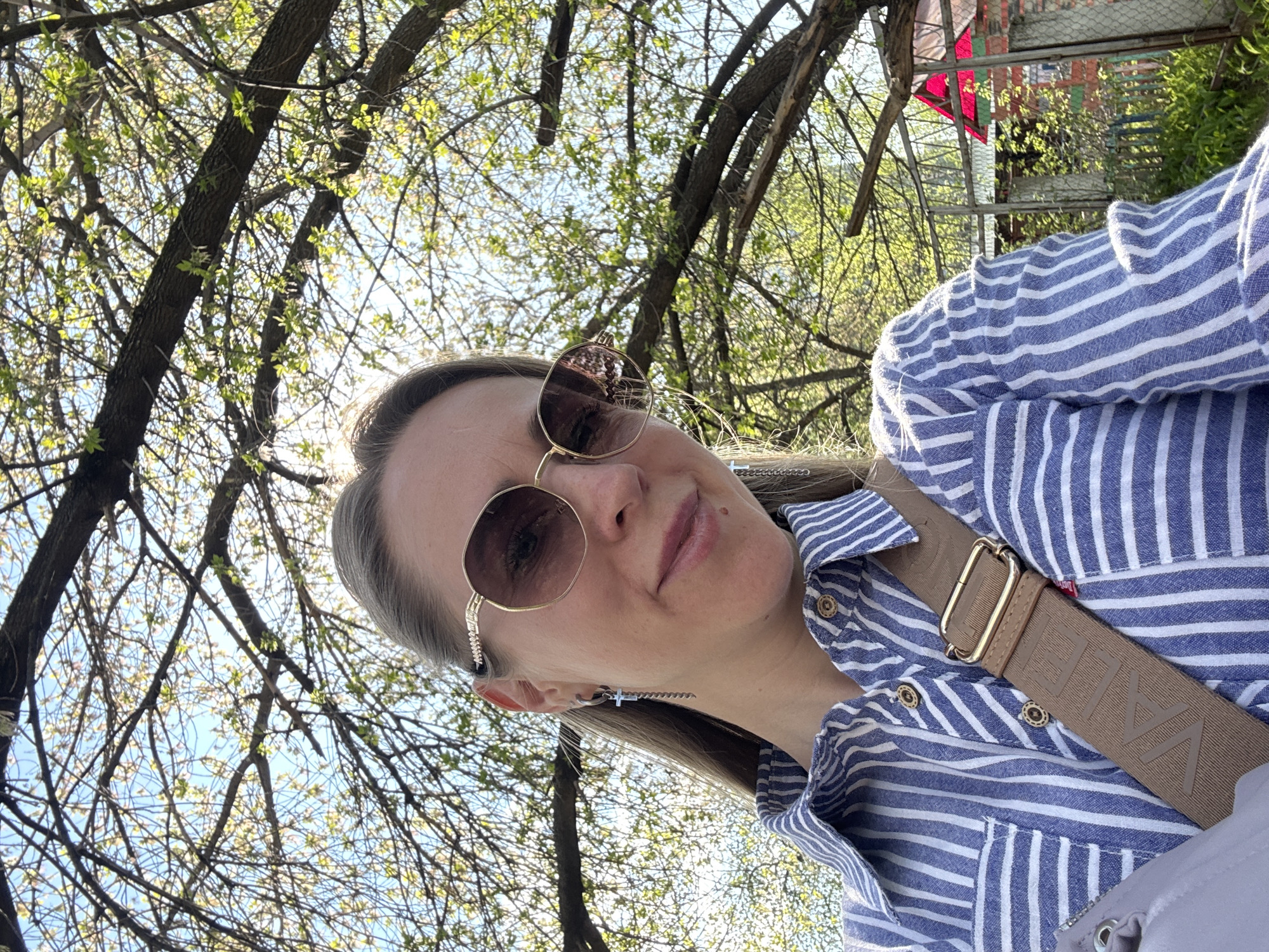 A fair-skinned woman with light brown hair, wearing round, gold-rimmed sunglasses, a blue and white striped shirt, and a tan-colored strap across her chest, is visible in the image. She appears to be outdoors under a tree with budding leaves. The background shows glimpses of a structure with red and green elements, likely within a park setting. Based on the prompt, the location is indicated to be in the city of Chaikovsky, Russia.
