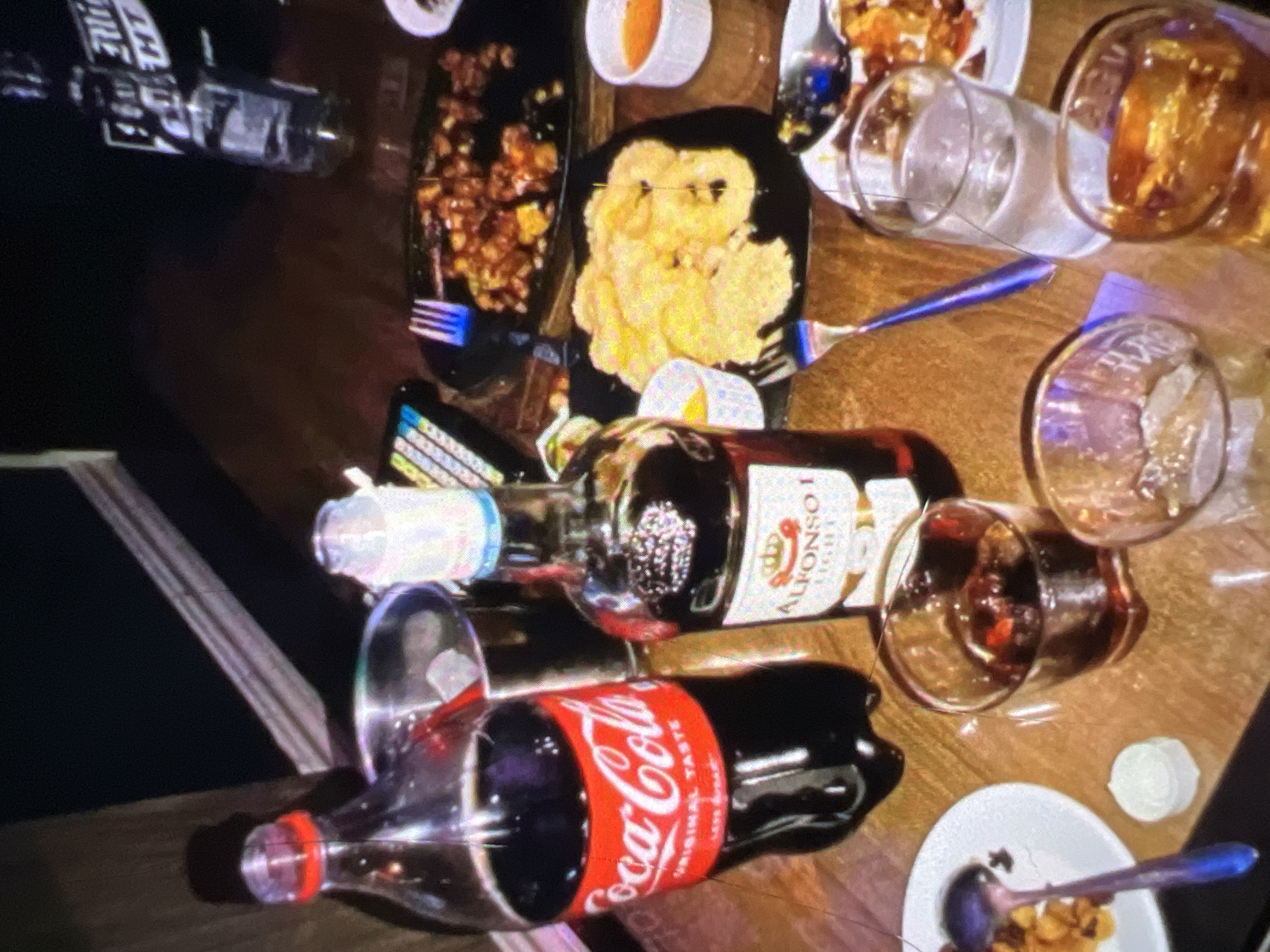 A close-up, high-angle shot depicts a glass-topped table laden with food and drinks, likely in a dimly lit restaurant or home setting in Lagos, Nigeria. The items on the table suggest a casual meal or gathering. Dominating the left side of the frame is a large, clear plastic bottle of Coca-Cola, its red and white label clearly visible. Next to it, to the right, is a bottle of Alfonso I brandy, identifiable by its label. Between these two bottles is a stainless steel ice bucket containing ice and what appears to be a straw.
Further to the right, on black serving platters, are portions of food. One platter holds what looks like crispy fried calamari rings, while another contains a savory dish, possibly a meat or vegetable stir-fry. Small white dipping bowls with orange sauce are also present.
Scattered across the table are several drinking glasses. One glass, positioned to the right of the brandy bottle, contains a dark liquid, likely brandy and ice. Another glass, closer to the foreground, is partially filled with a similar dark liquid. A third glass, to the right, is filled with clear water and ice. A fourth, empty glass sits in the foreground.
Cutlery, including a fork with a blue handle and a spoon with a blue handle, are visible among the dishes. A small, white, circular container, possibly for a condiment or sauce, is situated near a plate on the left. In the background, a partially visible bottle with a white label and a dark liquid stands upright. The overall impression is one of a shared meal or social occasion, with multiple individuals present but not directly visible in the frame. The lighting is dim, with some reflections on the glass table and bottles. The surface of the table appears to be a warm-toned wood or laminate.