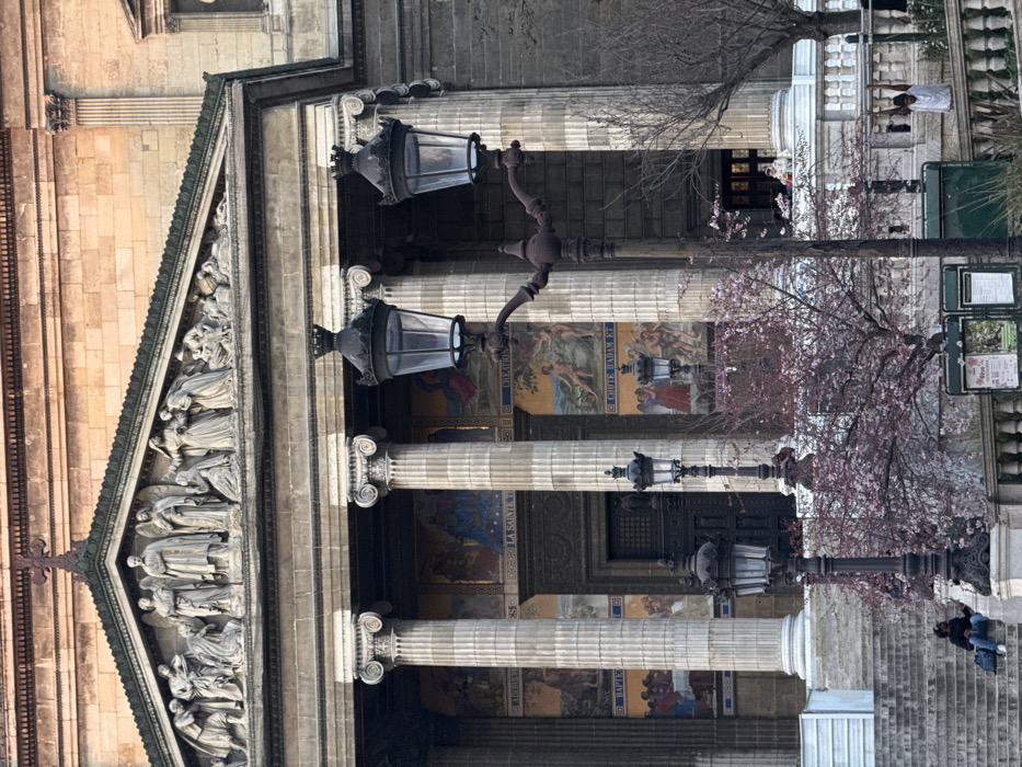 The image displays the neoclassical facade of Église de la Madeleine (Church of Saint Mary Magdalene) in Paris, France. The upper portion of the building features a large triangular pediment containing a detailed high-relief sculpture depicting numerous figures. Below this, an entablature is supported by a colonnade of tall, fluted columns with ornate capitals. Between the columns, several colorful mosaic panels illustrating biblical scenes are visible, including one titled "LA SAINTE TRINITÉ" and another "CHUTE D'ADAM ET..." (Fall of Adam and...). The central entrance, appearing as a dark, recessed doorway, is situated behind the columns.
In the mid-ground, a prominent, dark-colored, ornate cast-iron street lamp with two lanterns extends from the right side of the frame. To its left, a similar, single-lantern street lamp is partially visible. A small deciduous tree with sparse pink blossoms, suggesting early spring, is positioned between these lamps. A stone balustrade runs horizontally across the lower mid-ground, behind the street lamps and tree.
On the wide stone steps leading up to the church on the far left, a single individual wearing dark clothing is seated, with a light-colored bag beside them. On the right side, behind the balustrade, another person in a light-colored top is partially visible. Further back, near the church's entrance, faint figures are present on the balustrade and within the shadowed portico. The building's stone facade is bathed in sunlight on its upper sections, while the colonnade and lower areas are in shadow. A dark green public information board is partially visible at the bottom center, behind the balustrade.