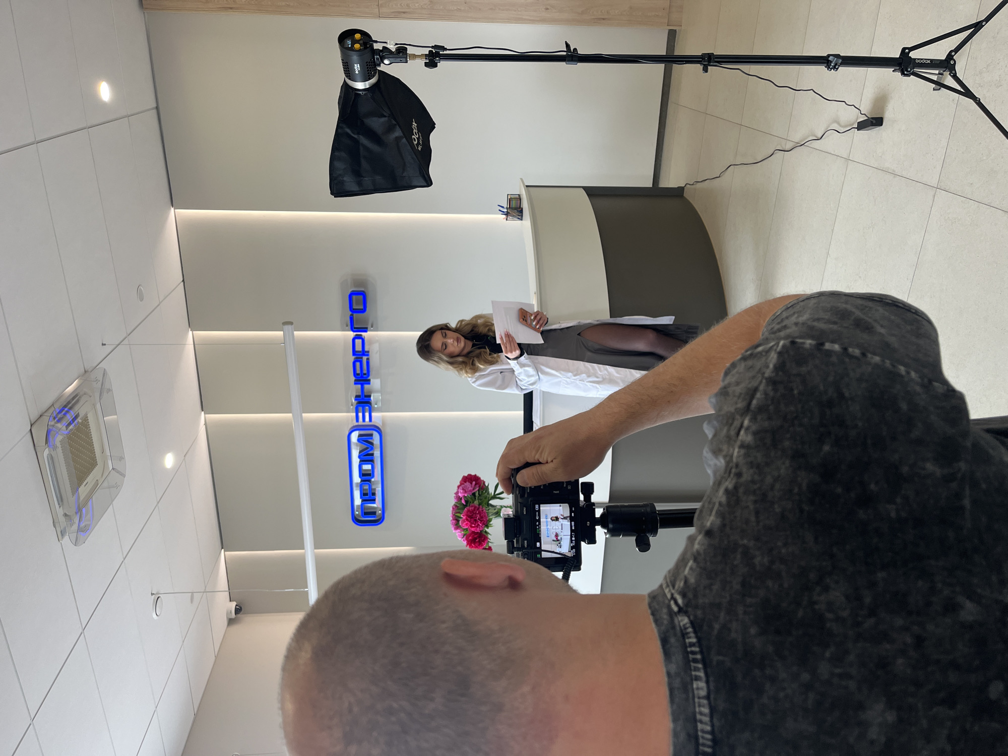 An indoor scene in Zelenodol'sk, Russia, captures a video or photoshoot in progress. In the immediate foreground, a person with a shaved head and wearing a dark, textured t-shirt is seen from behind, operating a digital camera mounted on a tripod. The camera's screen, displaying the live view, is visible.
In the midground, a woman with long, wavy blonde hair stands in front of a modern reception desk. She is wearing a white lab coat over a dark-colored dress or skirt with a high slit, and holds several white papers in her hands. She appears to be presenting or posing for the camera.
The background features a light-colored wall with a prominent blue illuminated sign displaying Cyrillic text: "ПРОМ ЭНЕРГО" (PROM ENERGO). To the right of the frame, lighting equipment is set up, including a "Godox" brand studio light with a softbox mounted on a tall stand. A power cable from the light runs across the light-colored tiled floor.
The ceiling above features white grid tiles, recessed circular lights, and a square air conditioning vent with blue internal lighting. A security camera is also visible mounted on the ceiling. On the reception desk, a small vase holds a bouquet of pink flowers, and a pen holder is also present. The overall setting suggests a professional environment, possibly a corporate office or medical facility, with a clean and contemporary design. The activity clearly indicates a planned media production.
