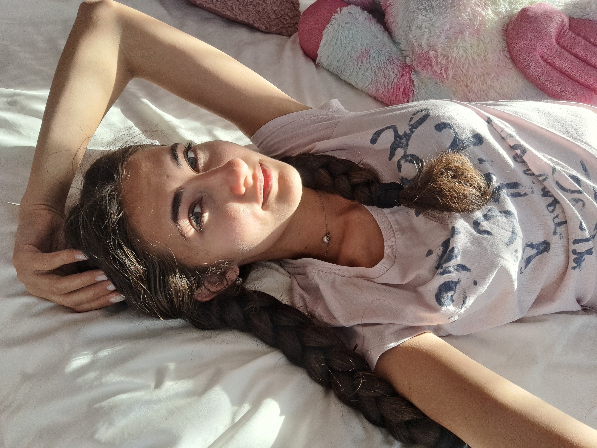A young woman lies on a white bed, facing the camera with her head resting on her hand. She is wearing a light pink T-shirt with a printed design. Her dark hair is styled in two braids that fall over her shoulders and onto the bed. Behind her, there is a pink pillow and a stuffed animal. The scene is bathed in sunlight. Location: Unknown, Unknown.