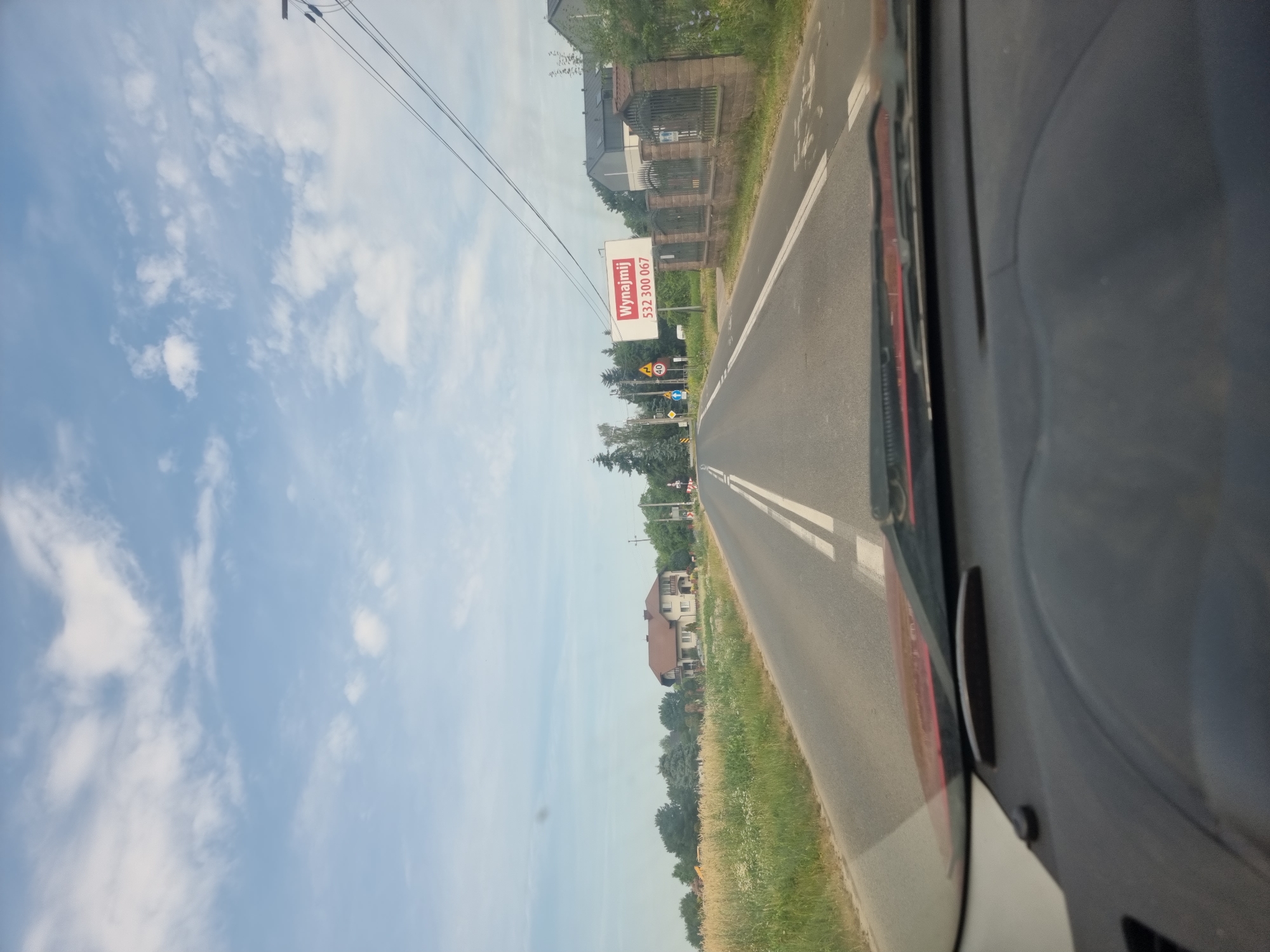 The image is taken from the perspective of someone driving a vehicle on a paved road. The road has a double white line down the center and appears to be in a rural or suburban area. To the left of the road, there is a field of tall grass and wildflowers, and further in the distance, a large, light-colored house with a brown roof. To the right of the road, there is a low stone fence with decorative metalwork, and behind it, a modern building with dark siding. A billboard is visible above the fence, featuring red text that reads "Wynajmij" and a phone number "532 300 067". Traffic signs, including a speed limit sign for 40, are also present along the roadside. The sky is blue with scattered white clouds. The image captures a daytime scene. No people are visible in the image.