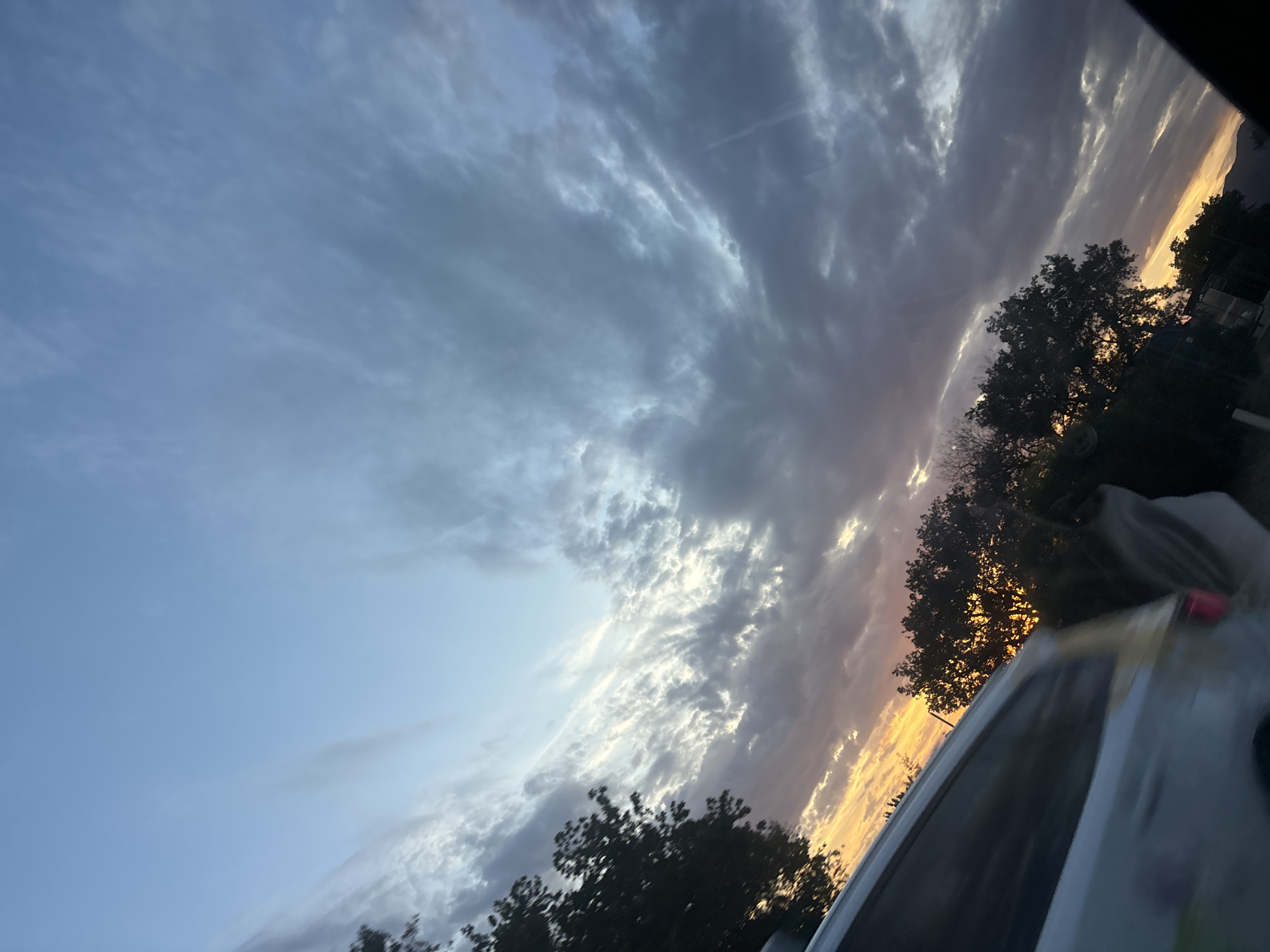 The image depicts a sunset or sunrise scene over Blizikuće, Montenegro. The sky, angled upwards from left to right, transitions from a clear, light blue at the upper left to vibrant yellows and oranges at the horizon, indicating a low sun position. A significant portion of the sky is covered by clouds: high, wispy cirrus clouds are visible against the bluer sections, while denser, darker cumulus or stratocumulus clouds dominate the right side of the frame and form a band above the horizon. These darker clouds exhibit tones of grey, purple, and blue, with patches illuminated by the low sun, creating lighter grey and white highlights.
Below the horizon, a dense line of trees, appearing as dark silhouettes, spans the lower mid-ground. Their dark foliage suggests deciduous trees. On the far right, a dark, indistinct landmass or distant hills are visible at the very bottom of the frame. In the lower left quadrant, a portion of a white or light-colored vehicle is visible, exhibiting motion blur consistent with being photographed from a moving perspective. A yellow or tan stripe runs horizontally along its side. The dark, curved shape on the extreme right edge of the image suggests the interior frame or window of the vehicle from which the photograph was captured. No individuals are present in the scene. The overall environment is an outdoor setting, likely rural or semi-urban, with natural foliage prominent against the dramatic sky.