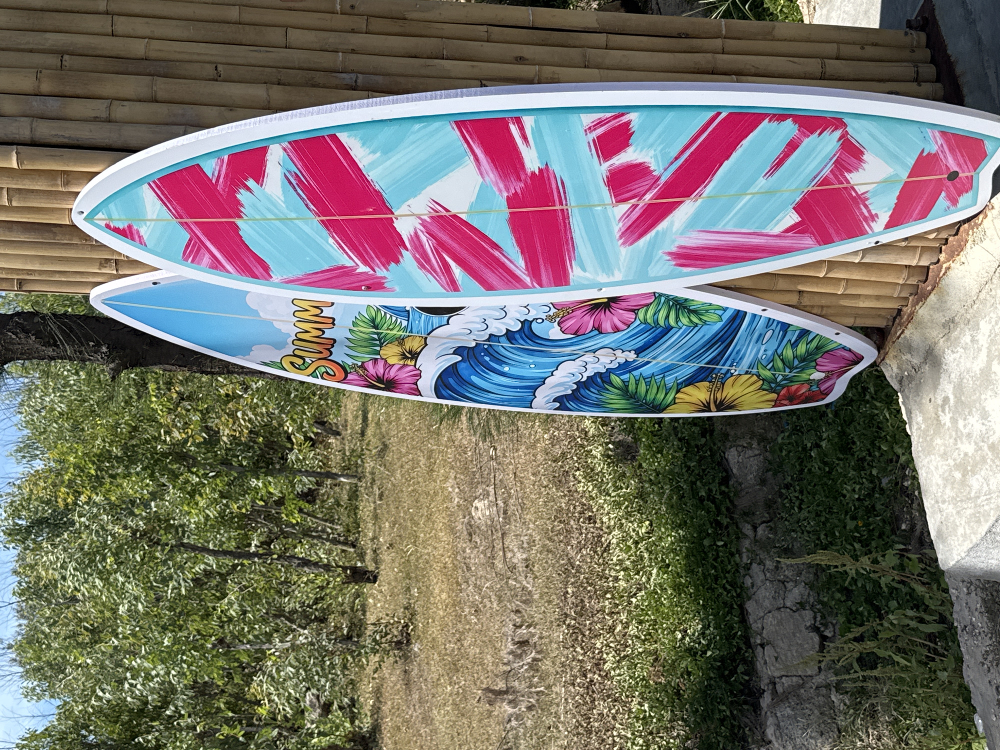 Two surfboards are propped against a bamboo wall. The surfboard on the left features a tropical wave design with the word "SUMM" and flowers. The surfboard on the right has a vibrant abstract design of pink and turquoise brushstrokes. The background shows a grassy area with trees. The scene is outdoors, likely in a coastal or recreational area. The location is identified as Phú Hội, Vietnam.