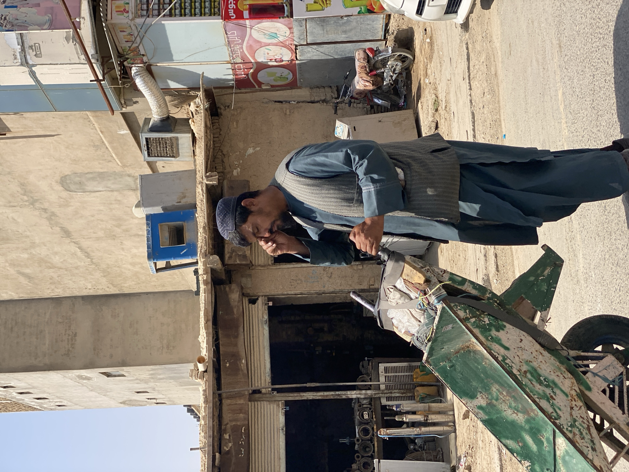 A man in a dark blue shalwar kameez and a gray vest, wearing a dark blue cap, stands by a rusty green pushcart. He appears to be rubbing his eye with his right hand. The pushcart is laden with various items, including what appears to be fabric or sacks, rope, and a wooden plank. Behind him, a storefront with corrugated metal doors is visible, with shelves filled with items such as water pumps and yellow containers. In the background, a red and white motorcycle is parked, and a white car is partially visible on the right. The buildings in the background are made of concrete and appear to be weathered. The overall scene depicts a street in Kandahar, Afghanistan, with a marketplace or workshop environment.