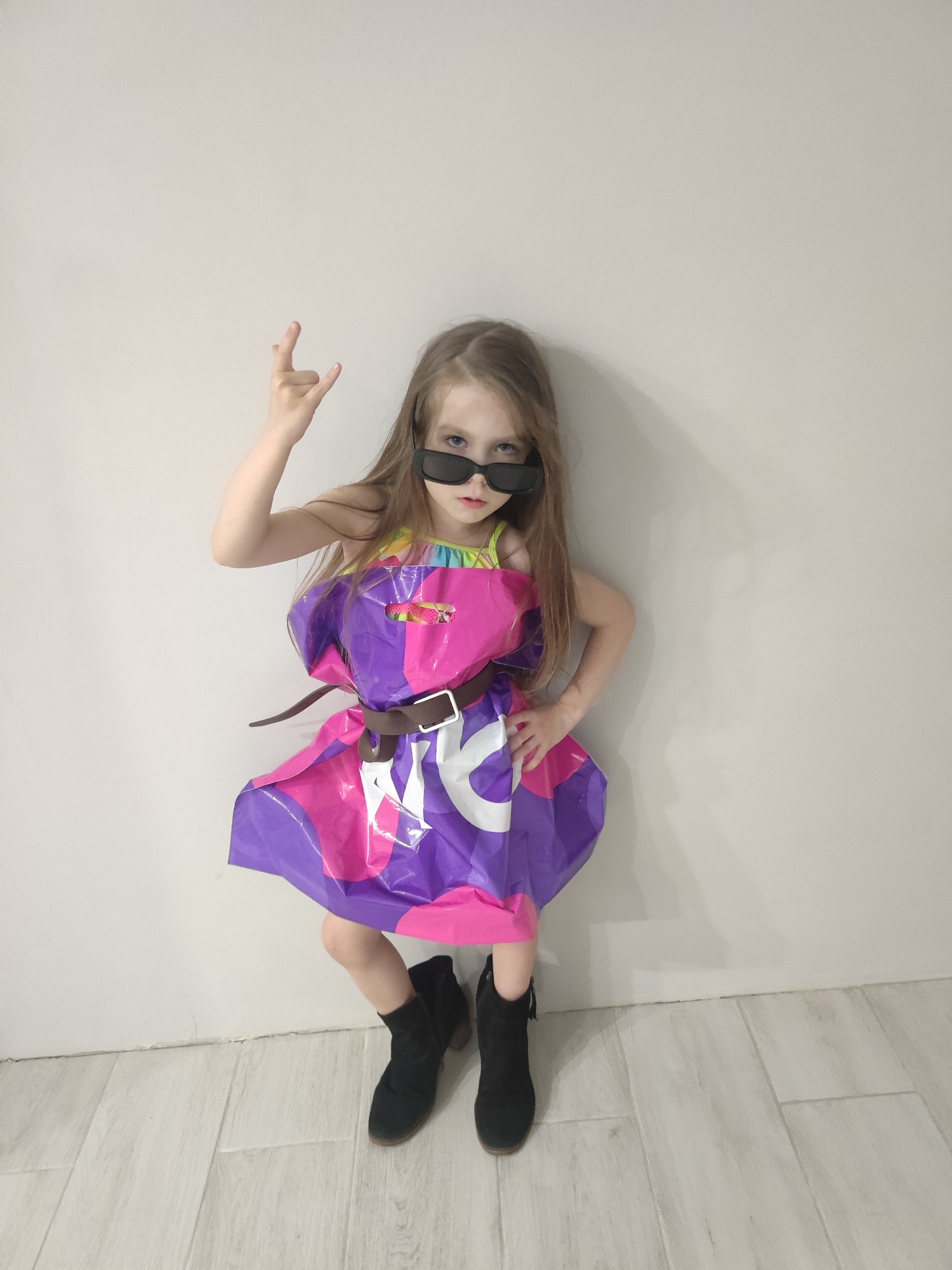 A young girl stands against a plain, light-colored wall. She is wearing black sunglasses and a dress made from a pink and purple plastic bag, cinched at the waist with a brown belt. She wears black ankle boots. Her right hand is raised, forming a "rock on" gesture. The location is potentially Votkinsk, Russia, though there is no visual evidence within the image to confirm this.