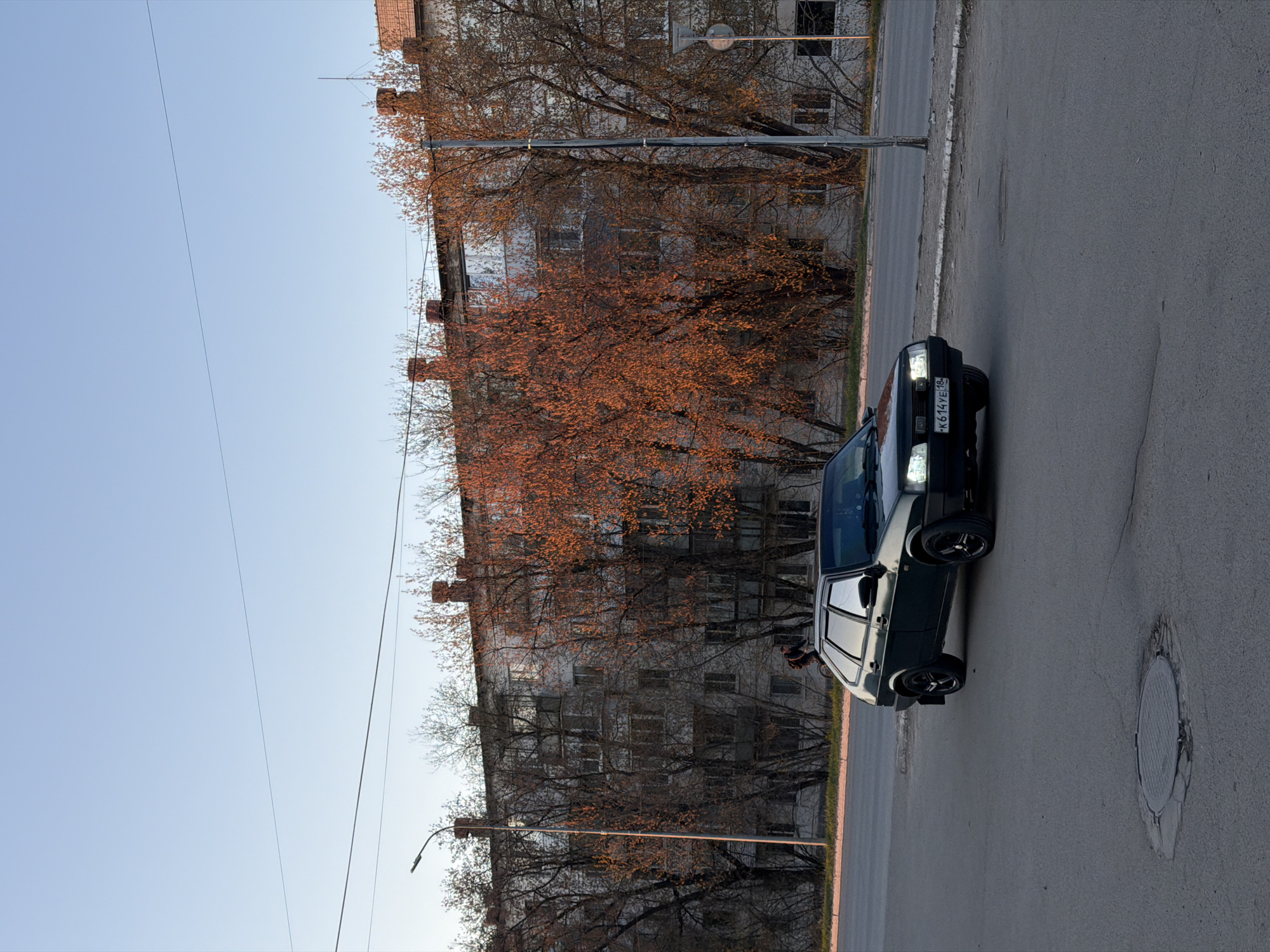 A black sedan, displaying license plate "K 614 YE/18" is parked on a gray asphalt road. The car's headlights are on. Behind the car, there is an old, six-story apartment building with many windows and chimneys on the roof. Bare trees with hints of orange leaves are in front of the building. The sky is blue. The scene is located in the city of Chaikovsky, Russia.