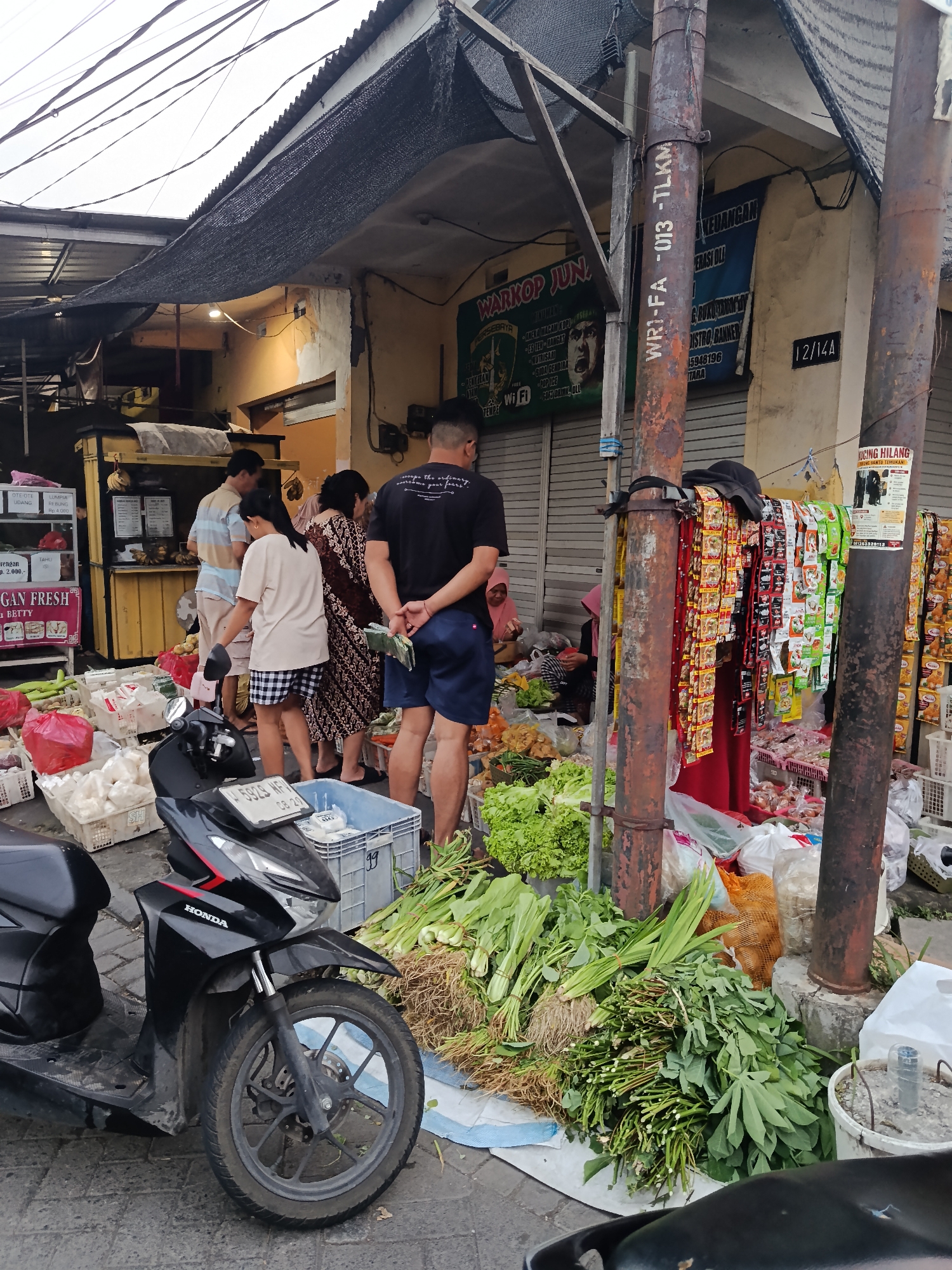The image depicts an outdoor market scene in Keboansikep, Indonesia. In the foreground, a black Honda scooter is parked on a paved surface. To the right of the scooter, a variety of fresh green vegetables, including leafy greens and root vegetables, are piled on the ground and on a white cloth. Behind the vegetables, a large metal pole stands upright, adorned with numerous small, colorful packets hanging from it. Further back, several people are visible. One person in a black t-shirt and blue shorts stands facing away from the camera. To their left, a woman in a patterned dress is interacting with another person. Behind them, a food stall with a yellow counter is set up. The market is covered by a black mesh awning supported by a metal structure. The environment appears to be a bustling street market with stalls and produce arranged for sale.