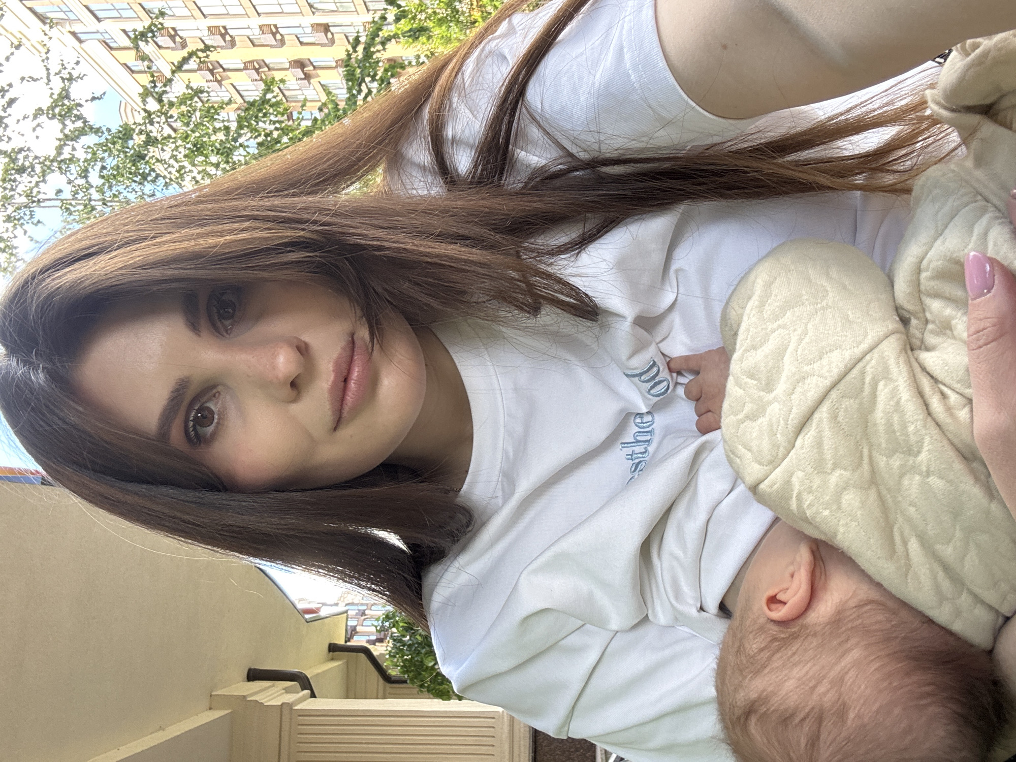 A woman with long, brown hair is breastfeeding a baby outdoors. The woman is wearing a white t-shirt and has light pink nail polish. The baby's head is visible, and it is partially covered by a beige blanket. The environment appears to be a residential area, with buildings and trees in the background. The location is Sofiivska Borshchahivka, Ukraine.