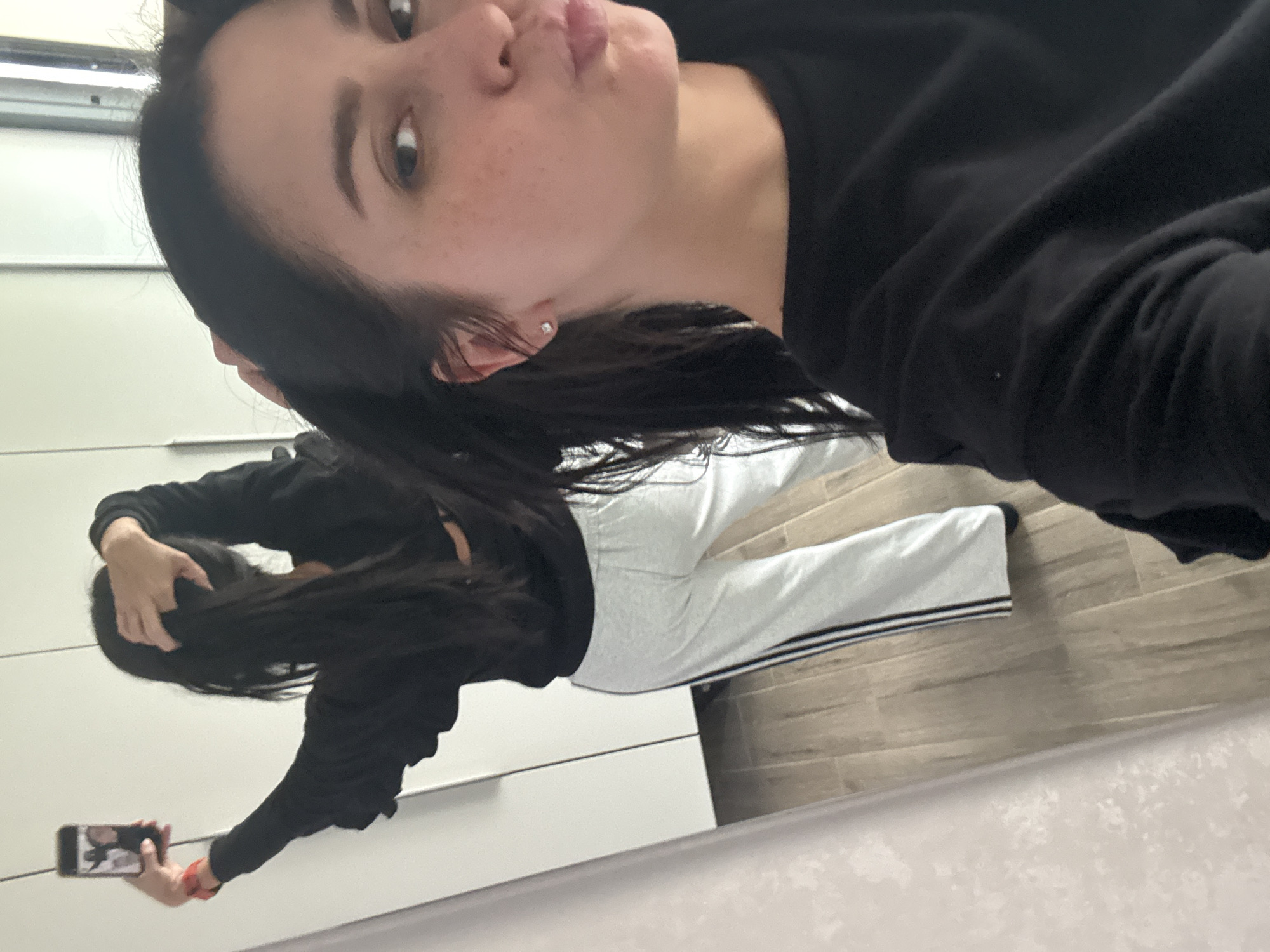 A young woman with long dark hair is taking a mirror selfie in what appears to be a bedroom or dressing area. She is wearing a black long-sleeved shirt and light grey sweatpants with black stripes down the sides. Her hair is partially pulled up, and she is holding a mobile phone in her left hand. The background shows a large white closet with mirrored doors and a wooden floor. The overall lighting is bright and appears to be natural or from overhead fixtures.