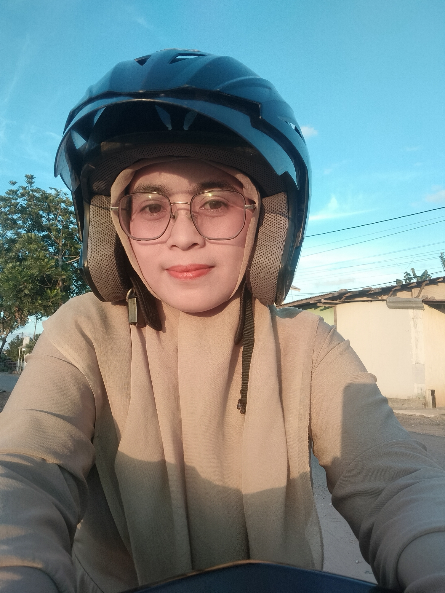 A selfie is taken by a woman wearing a beige hijab, glasses, and a black helmet. She is looking directly at the camera with a slight smile. She is wearing a loose-fitting beige top. The background shows a clear blue sky with some light clouds. To the left of the woman, there is a green tree. To the right, there is a building with a corrugated metal roof and a light-colored wall. The setting appears to be outdoors, possibly in a residential area of Karang Indah, Indonesia, given the architecture and vegetation. The lighting suggests it is either early morning or late afternoon. The woman is likely riding a motorcycle or scooter, as indicated by the helmet and the perspective of the photo.