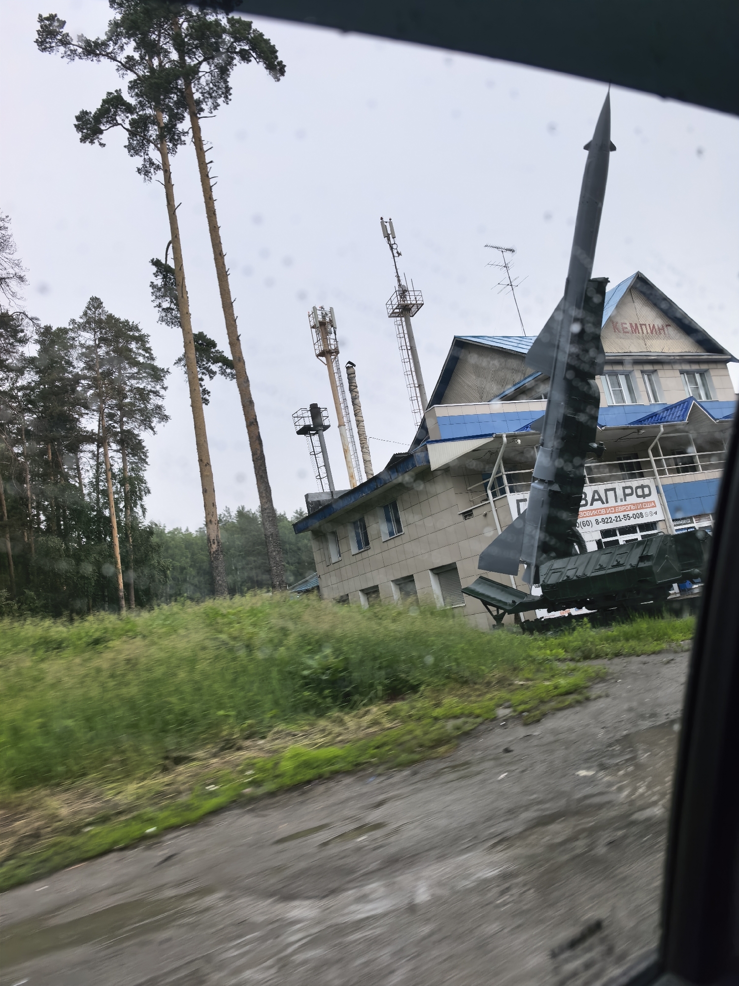 An outdoor shot captures a missile on display in front of a light-colored building with a blue roof. The missile is dark green and appears to be mounted on a trailer. The building has several windows and a sign that reads "ЗАП.РФ" and "КЕМПИНГ" in red letters. In the background, tall pine trees stand against a cloudy sky. The ground in front of the building is covered with green grass, and a wet, asphalt road is visible in the foreground. Several cell towers are also present in the background. The image is taken from inside a vehicle, as evidenced by reflections on the window.