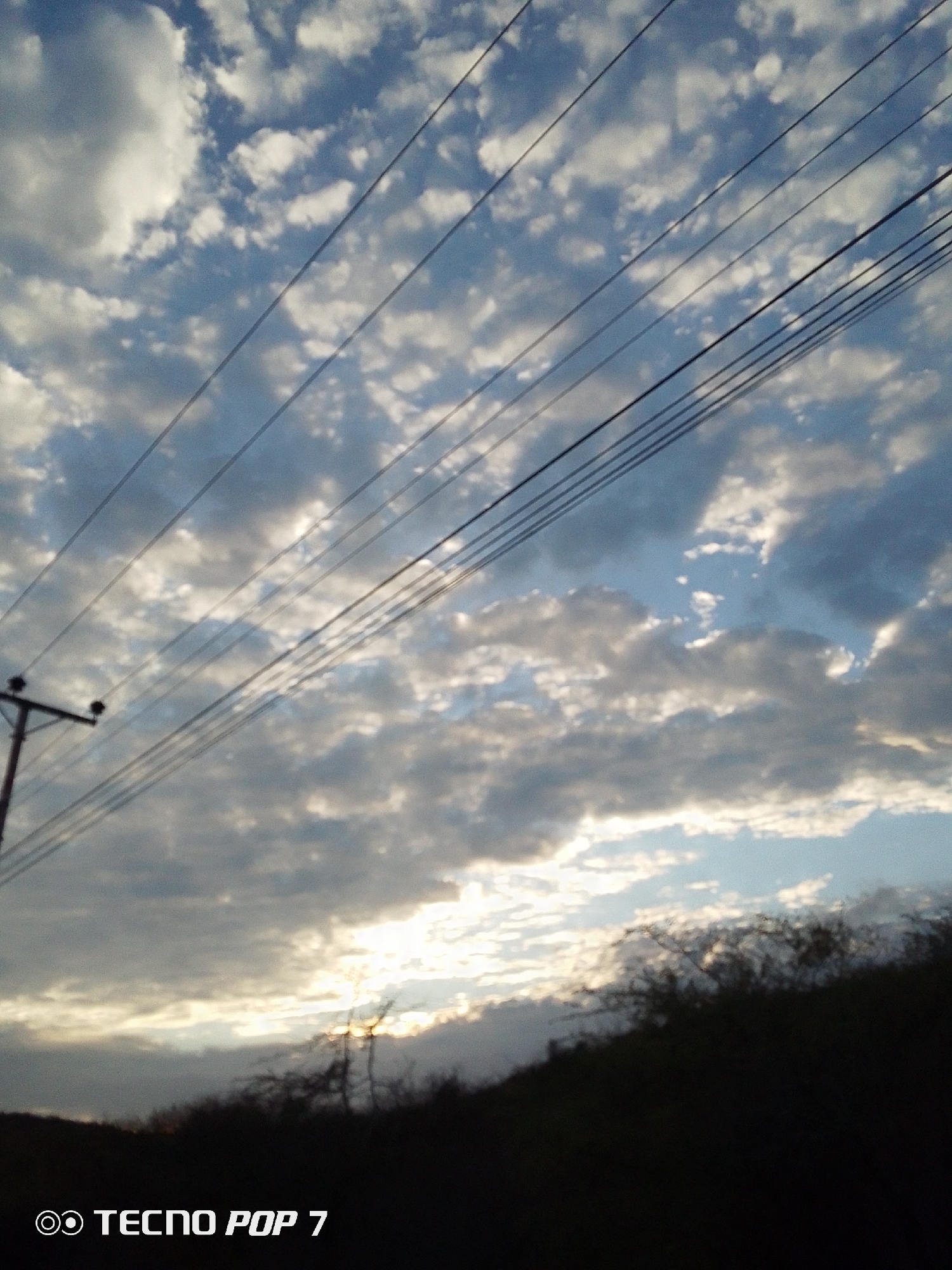 The image shows a view of the sky with numerous clouds, some partially obscuring the sun, over what appears to be a hill or ridge in Altagracia, Venezuela. Power lines stretch across the sky from a utility pole on the left. The foreground is dark, obscuring details of the ground and vegetation. "TECNO POP 7" is imprinted at the bottom.