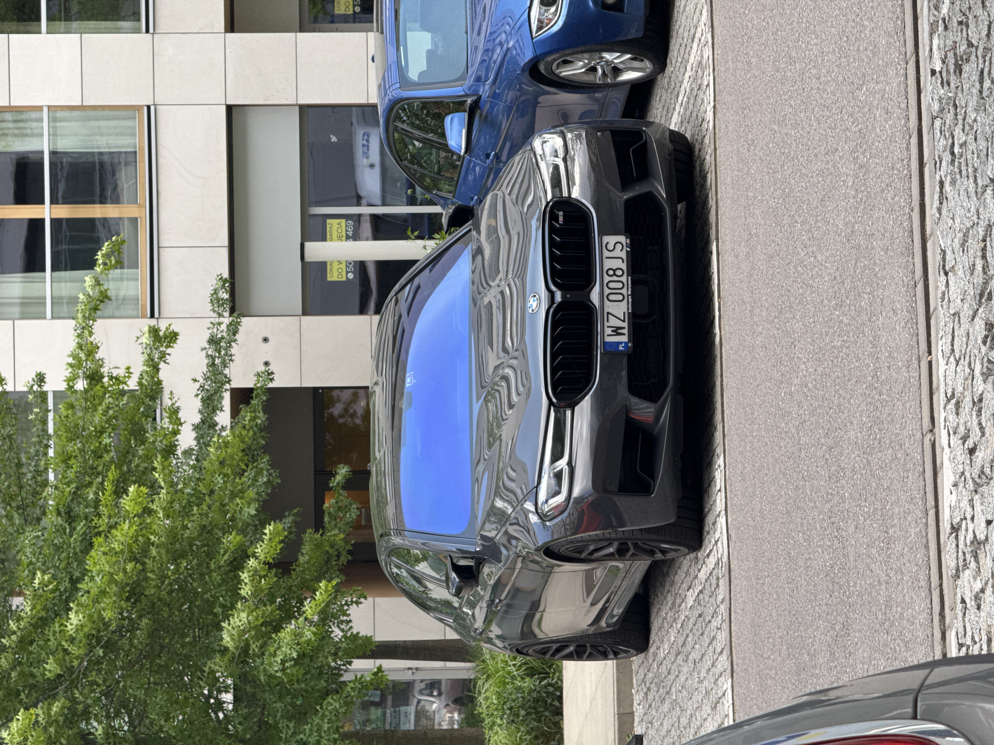 A dark grey BMW, likely an M5 model identifiable by the badge on its grille, is parked on a paved surface next to a modern building. A blue BMW is partially visible behind it. The dark grey car's windshield reflects a bright blue sky, indicating a sunny daytime setting.
The car is positioned alongside a sidewalk composed of both asphalt and cobblestones. To the left, a large green tree with dense foliage partially obscures the building. The building itself features a contemporary design with light-colored panels and rectangular windows, some of which reflect the surroundings.
Visible text on the dark grey BMW's front license plate reads "WZ 008 JS," with "PL" and "Inchcape Warszawa" also present, suggesting the location is in Poland, specifically near Warsaw. Several partially visible signs are affixed to the ground floor of the building, displaying text fragments such as "LOKAL DO W," "GARAZ JĘCIA," "4469," and "LOK DO." No people are visible in the scene.