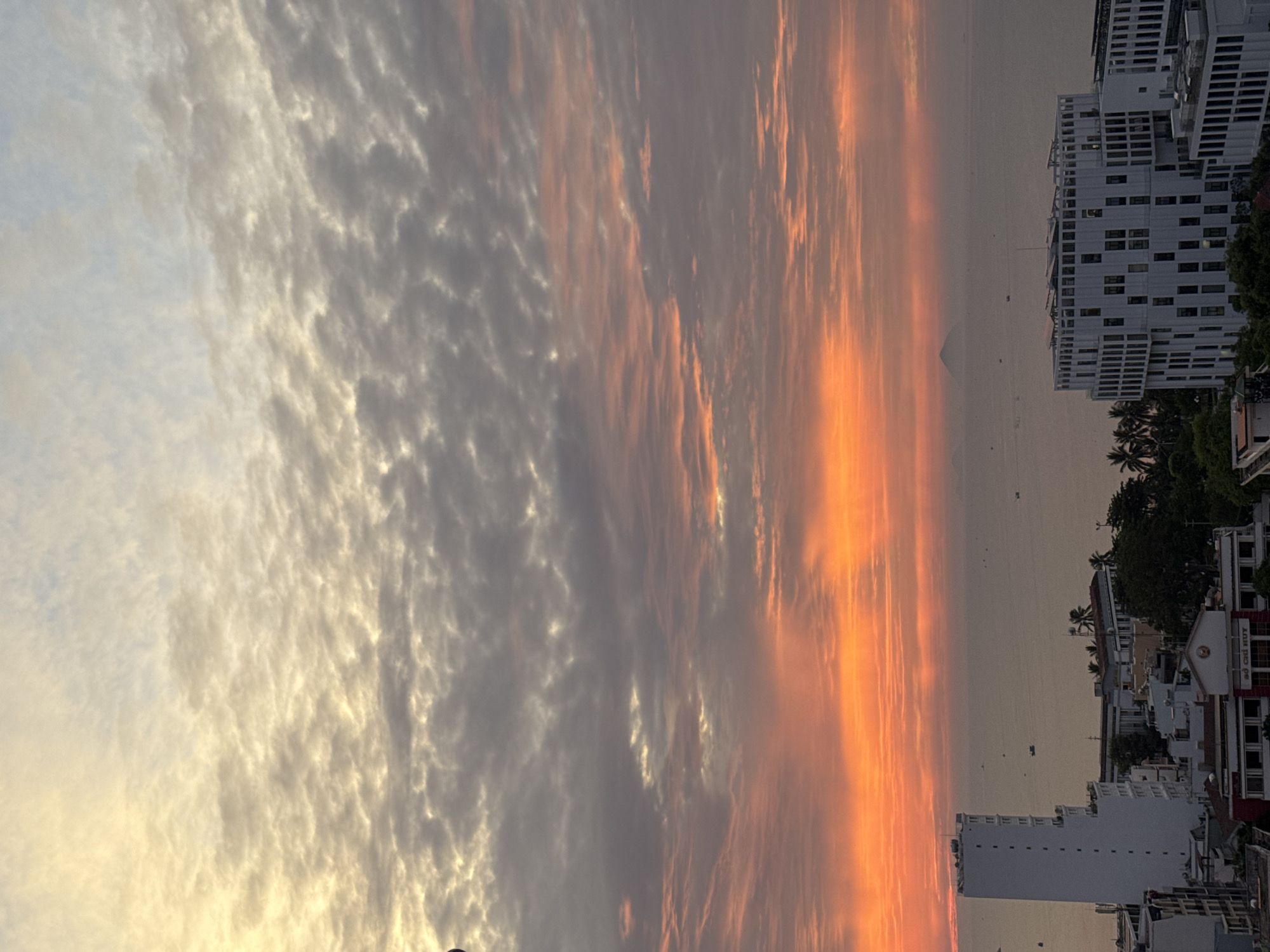 This image captures a sunset over the city of Nha Trang, Vietnam, seen from a high vantage point. The sky is ablaze with vibrant colors, transitioning from a bright orange at the horizon to shades of pink and gray as it reaches higher. Clouds are scattered across the sky, illuminated by the setting sun. In the foreground, several buildings of varying heights and architectural styles are visible. To the left, a tall, white, rectangular building stands out. To the right, a modern, multi-story building with a distinctive facade features numerous windows. The sea stretches out in the background, dotted with small boats. In the far distance, a triangular-shaped island or mountain is faintly visible on the horizon. The overall scene depicts a city skyline bathed in the warm glow of a sunset over the ocean.