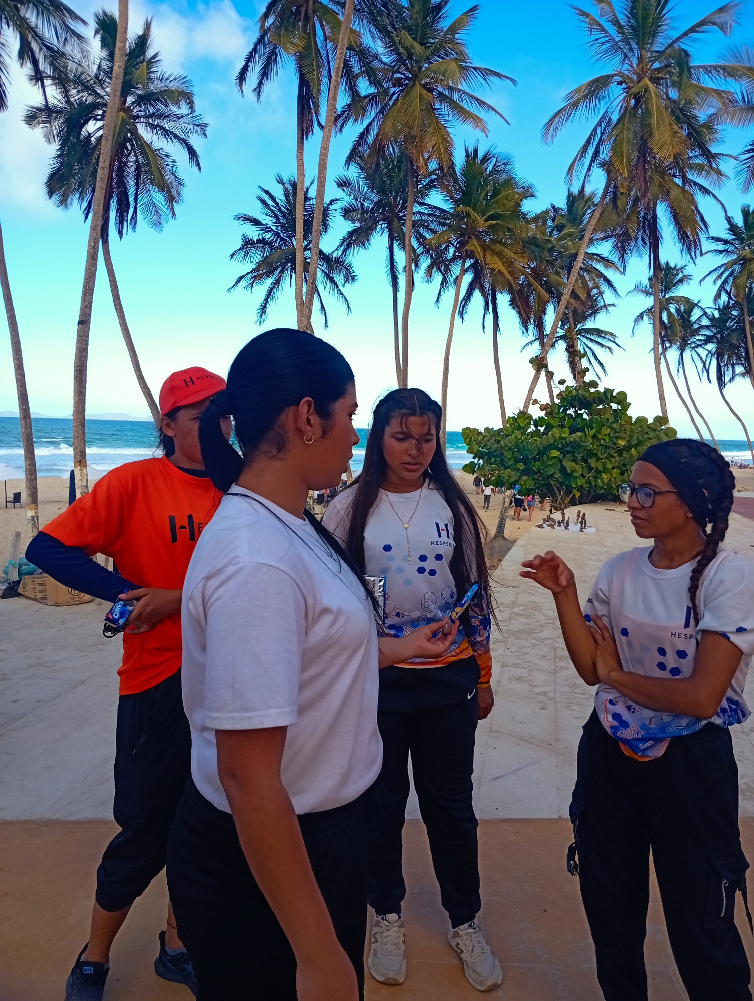 Four young women stand on a paved area, likely a walkway, at Playa El Agua, Venezuela. The sky is blue with scattered clouds, and numerous tall palm trees are visible in the background. The ocean and beach are visible in the distance.
The woman closest to the camera is facing left, wearing a white t-shirt and black pants. The woman to her left wears an orange shirt, black pants, and a red baseball cap. She holds a small item in her left hand. To the right of the first woman, another woman wears a white t-shirt with a logo, black pants, and sneakers. She is holding a blue object. The woman furthest to the right wears a similar white t-shirt, black pants, a black headscarf, and glasses. She has her arms crossed and is speaking.
In the background, people are walking on the beach near a small, dense group of shrubs. There are also small structures visible.
