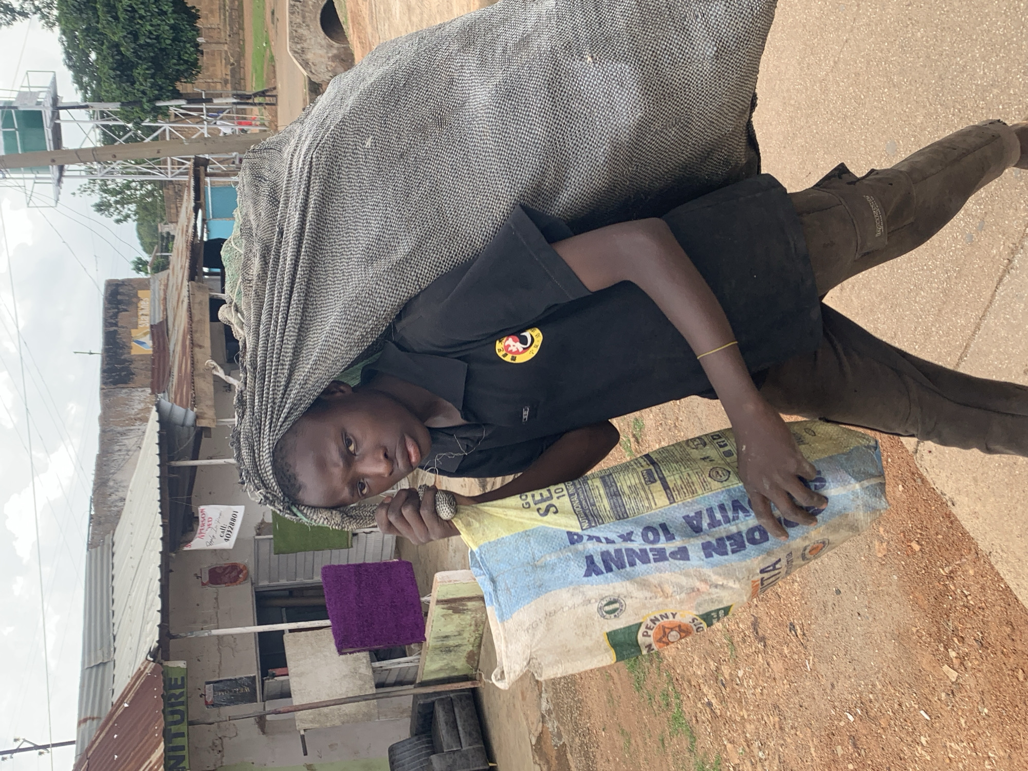In this medium shot, a young person is shown from the knees up, carrying a large, heavy load on their back, partially covered by a grey, textured tarp. The person is wearing a black t-shirt with a yellow and red emblem and dark pants. They are also wearing a gold bracelet on their left wrist. The load appears to be a large sack or bag, held in place by the tarp. In their hands, they are holding the bottom of a lighter-colored bag with blue and yellow text, which appears to be a food or grain sack, with the text "DEN PENNY VITA 10x" clearly visible. The background suggests an outdoor setting in a somewhat rustic environment. There are buildings with corrugated metal roofs, some with signs. One sign displays "FURNITURE" and another has the text "PERSON ED" and a phone number. In the distance, there's a water tower structure and some greenery. The ground is dusty and appears to be a dirt path or road. The lighting suggests it could be daytime, possibly overcast due to the diffuse light. The person's expression is neutral, focused on their task. The scene depicts the act of carrying a substantial load, likely for work or transport, in what appears to be a marketplace or residential area in Jos, Nigeria.