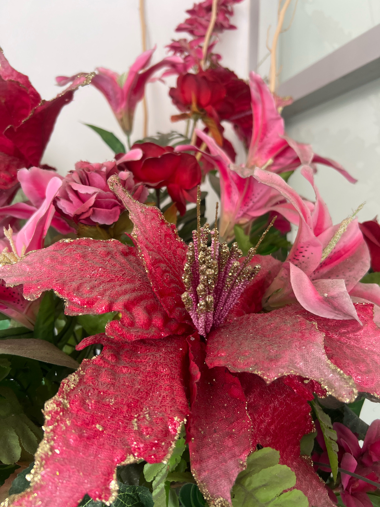 This is a close-up, eye-level, medium shot photograph of a festive artificial flower arrangement. The arrangement features various shades of pink and red flowers, with some appearing to be poinsettias and lilies. The petals of the flowers are textured and have a glittery gold trim, giving them a shimmering effect. Some of the flowers have pink stamen with gold glitter. The background is out of focus and appears to be a neutral, indoor setting with a white wall and a gray element that could be part of a window frame or furniture. There are no people or animals visible in the image, and no specific actions are taking place. The lighting is soft and diffused, highlighting the textures and sparkle of the artificial flowers. The photograph is cropped to focus on the details of the floral arrangement, with the arrangement likely positioned indoors in a city like Yogyakarta, Indonesia, given the context provided.