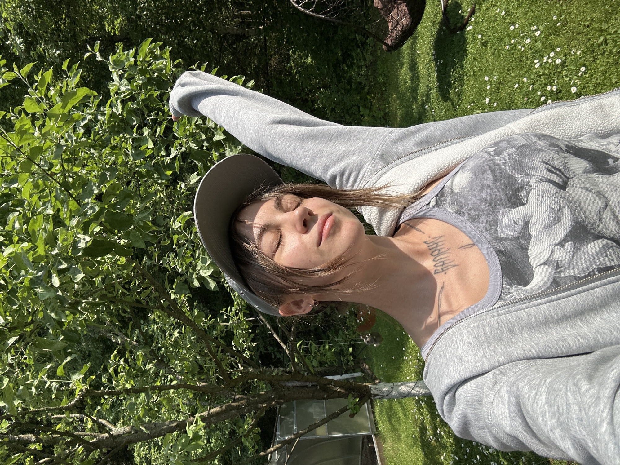 A young woman with brown hair, wearing a grey baseball cap, a grey zip-up hoodie, and a graphic tank top, stands in a grassy outdoor area. Her eyes are closed, and her right arm is raised above her head. She has tattoos on her neck. In the background, there is a small greenhouse, a tree, and dense green foliage. The ground is covered in green grass dotted with small white flowers. A dark, woven chair hangs from a stand in the background. The scene appears to be set in a yard or garden.