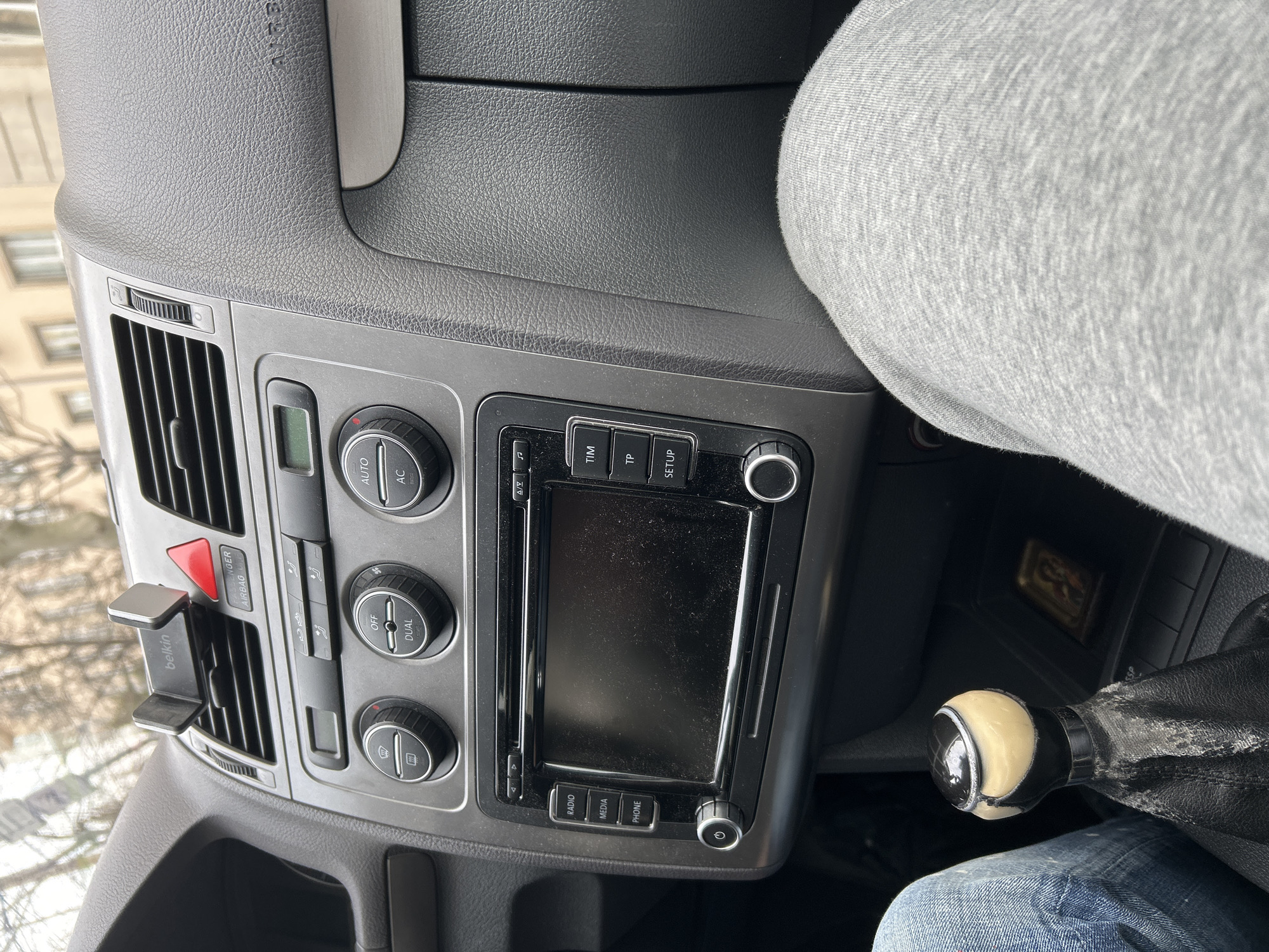 The image depicts the interior of a passenger vehicle, viewed from the front passenger seat looking towards the dashboard and front windshield. The dashboard is dark grey and textured. A "Belkin" brand phone holder is clipped into the central air vent. Below this, a red triangular hazard light button is visible. The center console features an infotainment system with a blank, dust-covered screen, flanked by buttons labeled "RADIO," "MEDIA," "PHONE" on the left, and "TIM," "TP," "SETUP" on the right. Below the screen are two rotary knobs. Above the infotainment system, climate control knobs and digital displays are visible, with a "DUAL" button present. The upper right dashboard area indicates "PASSENGER AIRBAG."
In the lower left foreground, the leg of a person wearing blue denim jeans is visible, consistent with a driver's position. In the lower right foreground, the leg of a person wearing light grey sweatpants or joggers is visible, consistent with a front passenger. Between the two visible legs, a manual transmission gear shifter is present, featuring a worn and discolored black leather boot and a light-colored knob with a dark top. Adjacent to the gear shifter, within a storage compartment, a small rectangular object resembling a religious icon or miniature picture frame is visible, faced upwards.
Through the front windshield, a blurred urban background is visible, showing light-colored building facades with multiple windows and bare tree branches, suggesting an overcast day. No specific landmarks or street signs are discernible to pinpoint a precise location within Sofia, Bulgaria from the visible background elements.