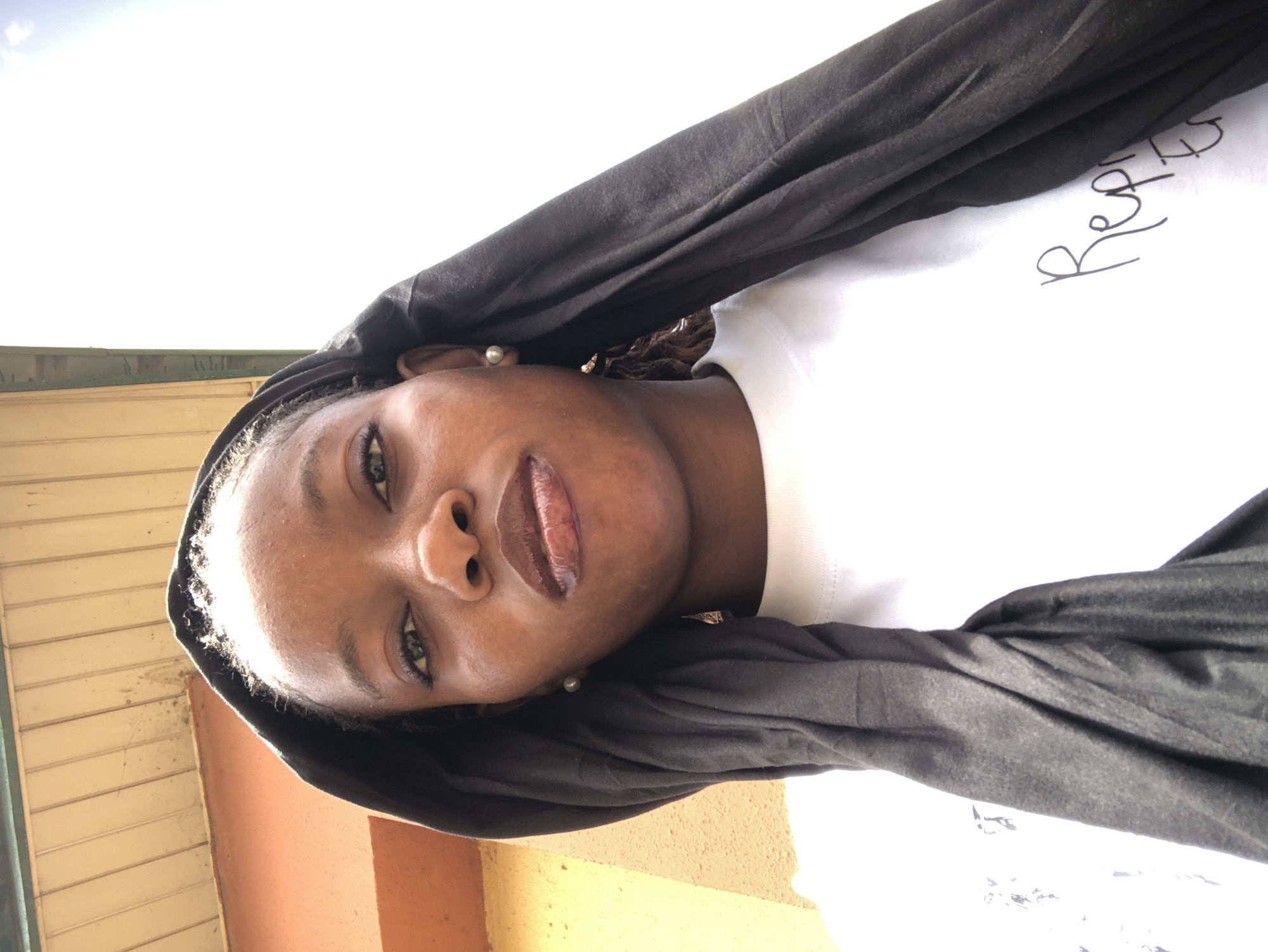 A woman with brown skin is facing the camera, head tilted slightly. She has dark hair with a black head covering. She is wearing a white t-shirt. Outside there is a yellow wall or side of a building. The location appears to be Pindiga, Nigeria.