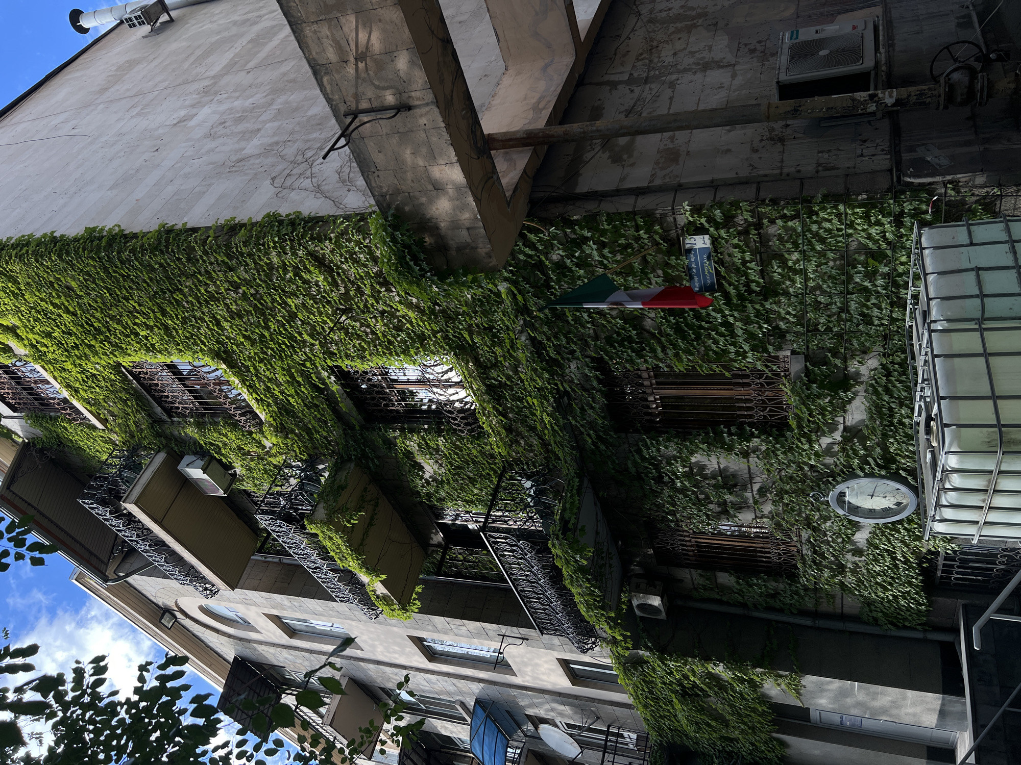 The original image, oriented sideways, depicts a tall building facade in Yerevan, Armenia, heavily covered in vibrant green ivy, shot from a low-angle perspective under a bright, clear blue sky with some white clouds.
The building's lower and middle sections are almost entirely engulfed by the dense ivy, creating a striking contrast with the architectural elements. Numerous windows, some with elaborate dark-colored wrought iron grates and others featuring balconies with similar ornate railings, are visible through the foliage. Several air conditioning units are mounted on the walls.
A prominent feature is a vertically striped flag (green, white, red) hanging from a pole near the center of the ivy-covered wall. Below the flag, a white rectangular plaque or sign is affixed, bearing text in Armenian script. This text is identifiable as "ԵՐԵՎԱՆԻ ԱԶԳԱՅԻՆ ՊՈԼԻՏԵԽՆԻԿԱԿԱՆ ՀԱՄԱԼՍԱՐԱՆ," which translates to "Yerevan National Polytechnic University." Further down on the wall, a large, circular, vintage-style clock with a white face and dark rim adds a classic touch.
Above the ivy line, the building's upper facade is made of light-colored, possibly stone or tiled material, featuring a modern, cylindrical vent or chimney pipe extending upwards against the sky. On the right side of the frame, parts of an adjacent structure with dark, weathered surfaces and industrial elements are visible. Some tree leaves are also seen in the bottom left corner.
The scene captures an urban environment on a sunny day, showcasing a blend of nature and architecture, with no specific human activity or event taking place. The heavy ivy coverage gives the old building a distinctive, charming character.