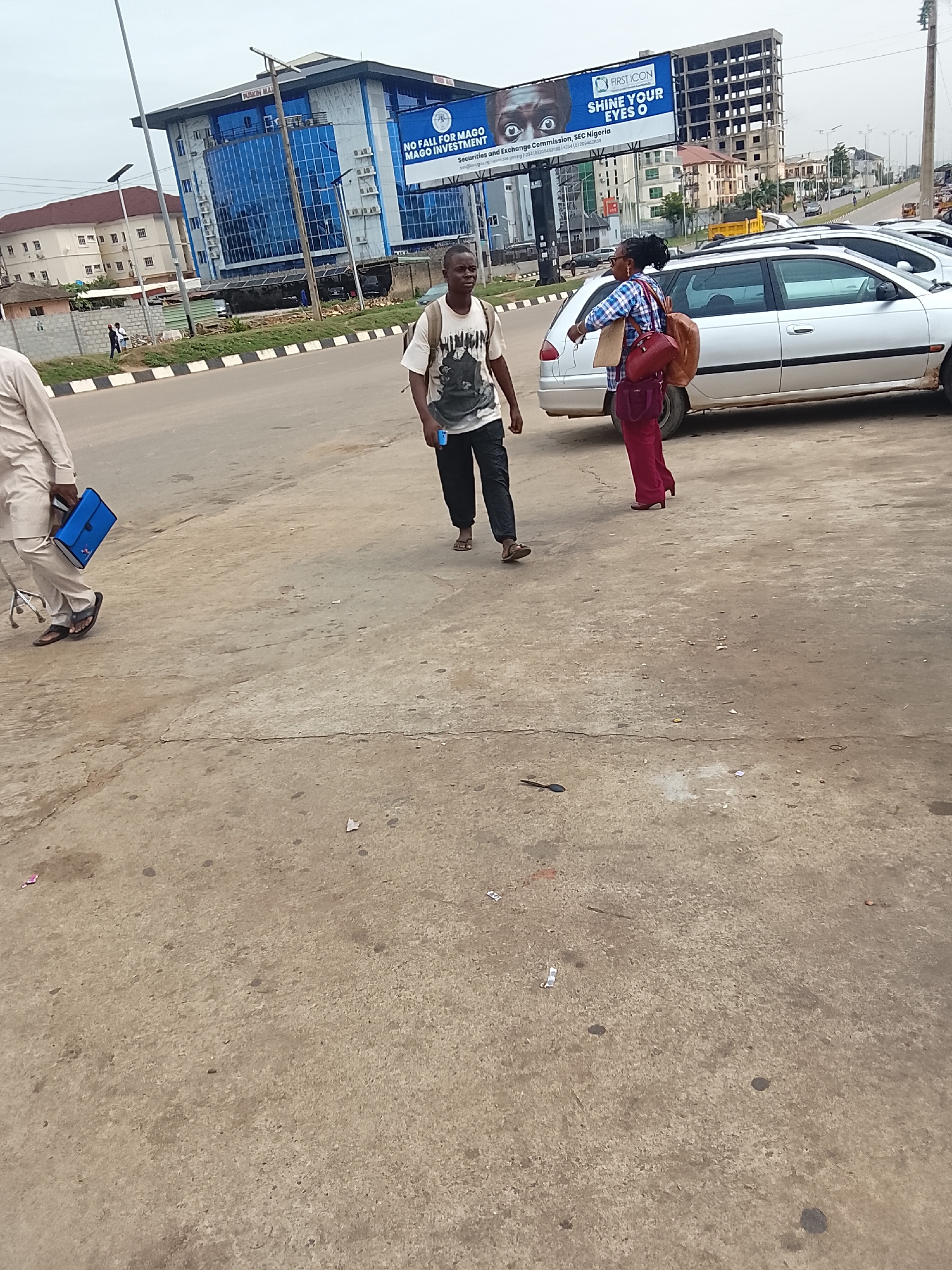 The scene is a street in Abuja, Nigeria, possibly near a business district, featuring a large billboard with an advertisement for "First Icon" and a warning against "Mago Investment" by the Securities and Exchange Commission (SEC) Nigeria. A modern glass-fronted building is visible behind the billboard, and an unfinished high-rise structure is in the background.
Several individuals are present. On the left, a man is walking while carrying a blue briefcase. In the center of the frame, another man is walking towards the right, wearing a t-shirt and carrying a bag. A woman is walking near a silver car, she is carrying a brown purse and papers.
The street appears to be a somewhat busy intersection. The pavement is concrete and shows signs of wear and stains.