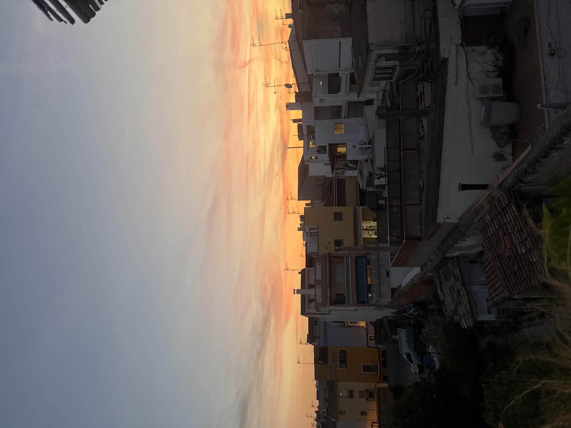 This is an eye-level, outdoor, full shot of a residential cityscape at sunset in Vilanova i la Geltrú, Spain. The sky is a gradient of light blue at the top, transitioning to soft orange and pink hues as it nears the horizon, with scattered wispy clouds. The foreground features a cluster of multi-story buildings, predominantly white and yellow, with tiled roofs. Many buildings have antennas on their rooftops. Some windows are illuminated, suggesting occupancy. In the middle ground, a dark silhouette of a cat is visible on a white wall. To the left, a dark green van is parked near some overgrown vegetation. The image captures a typical urban residential setting bathed in the warm light of twilight.