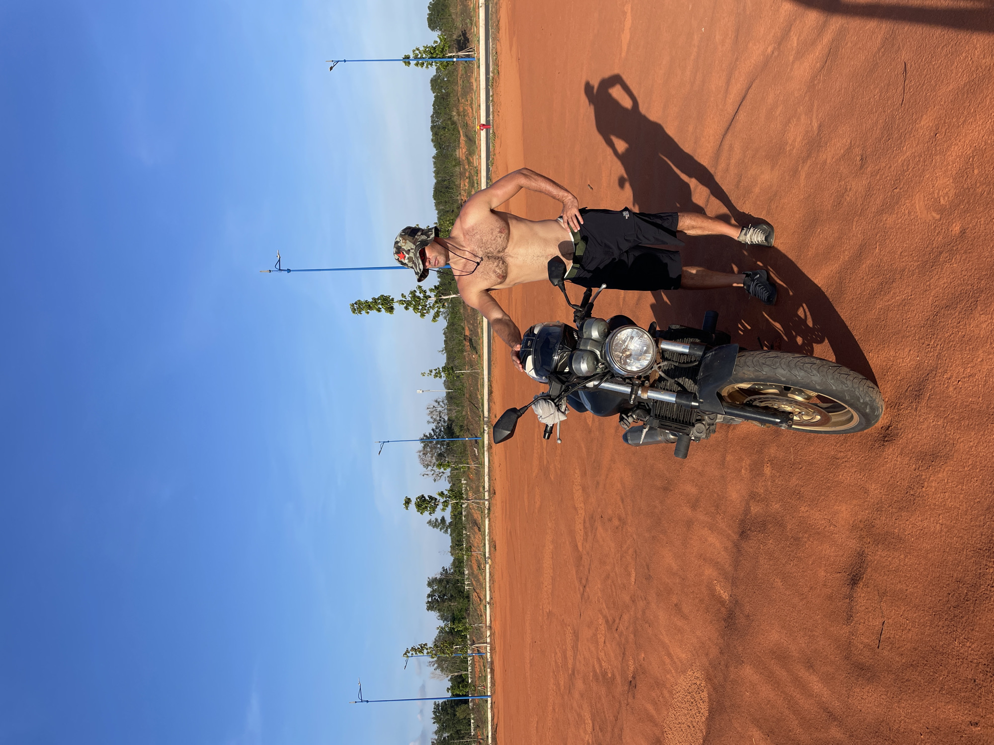 In Mũi Né, Vietnam, a shirtless man wearing a camouflage bucket hat and black shorts stands next to a black motorcycle on a large, orange-red dirt area under a clear blue sky. The man, with his hands on his hips and resting on the motorcycle, is looking to his left. The motorcycle is positioned in the foreground, facing left, with its headlight illuminated. In the background, there are several blue lampposts, sparse trees, and a distant line of vegetation. The sun casts a long shadow of the man and the motorcycle onto the dusty ground.