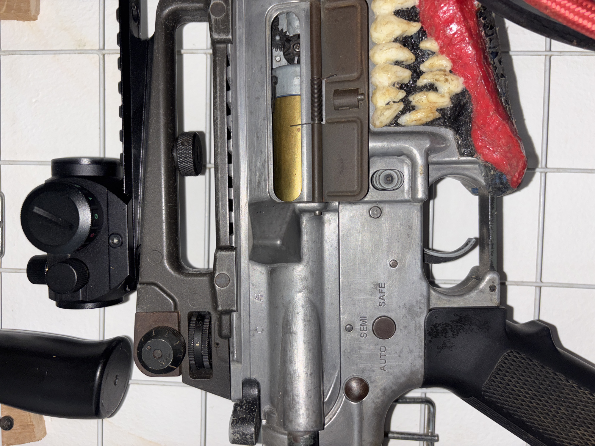 The image is a close-up, forensic-style photograph of the receiver and stock of a firearm, likely an assault rifle, modified with decorative elements resembling teeth and a red tongue. The firearm is mounted on a white grid background, possibly a wall or display.
Key details visible on the firearm include:
* **Optics:** A black red dot sight is mounted on the top rail.
* **Receiver:** The metallic receiver shows markings including "C", "A", "SEMI", "AUTO", and "SAFE", indicating fire selection controls. A gold-colored cartridge is visible within the ejection port.
* **Trigger group:** The trigger and safety selector are visible.
* **Grip:** A black textured pistol grip is attached to the lower receiver.
* **Decorative Modification:** The most prominent modification is a grotesque element resembling a monster's mouth, complete with yellowish teeth and a red tongue, affixed to the front of the receiver near the trigger guard.
The lighting is even, and the focus is sharp on the firearm, suggesting the image was taken intentionally to document the object. There are no people present in the image, and no actions are being performed.
This image does not provide any information that would allow for the determination of its location in the city of West Jakarta, Indonesia, or any other geographical location. The content is solely focused on the object itself and its immediate surroundings.