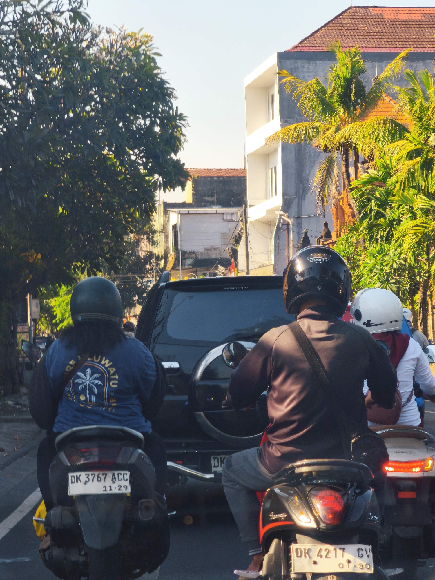 In Denpasar, Indonesia, on a street lined with trees and buildings, a traffic scene unfolds during daylight. Three motorcycles are visible, two in the foreground and one partially in the background. The first motorcycle, closest to the viewer, is ridden by a person wearing a dark helmet and a black long-sleeved shirt. Their back is to the camera, and they are riding a black scooter with a license plate reading "DK 3767 ACC" and "11.29". They wear a dark blue shirt with a graphic of a palm tree and text that reads "LUW ATU".
To the right of the first motorcycle, a second motorcycle carries two individuals. The rider wears a white helmet and a white long-sleeved shirt with a red garment underneath. The passenger behind them wears a red garment and a white helmet. Their scooter has a license plate reading "DK 4217 GV" and "01.30".
Between the two foreground motorcycles, a dark SUV is positioned. The environment is characterized by tropical vegetation, including palm trees, and buildings with red-tiled roofs and light-colored facades. The lighting suggests it is either morning or late afternoon.