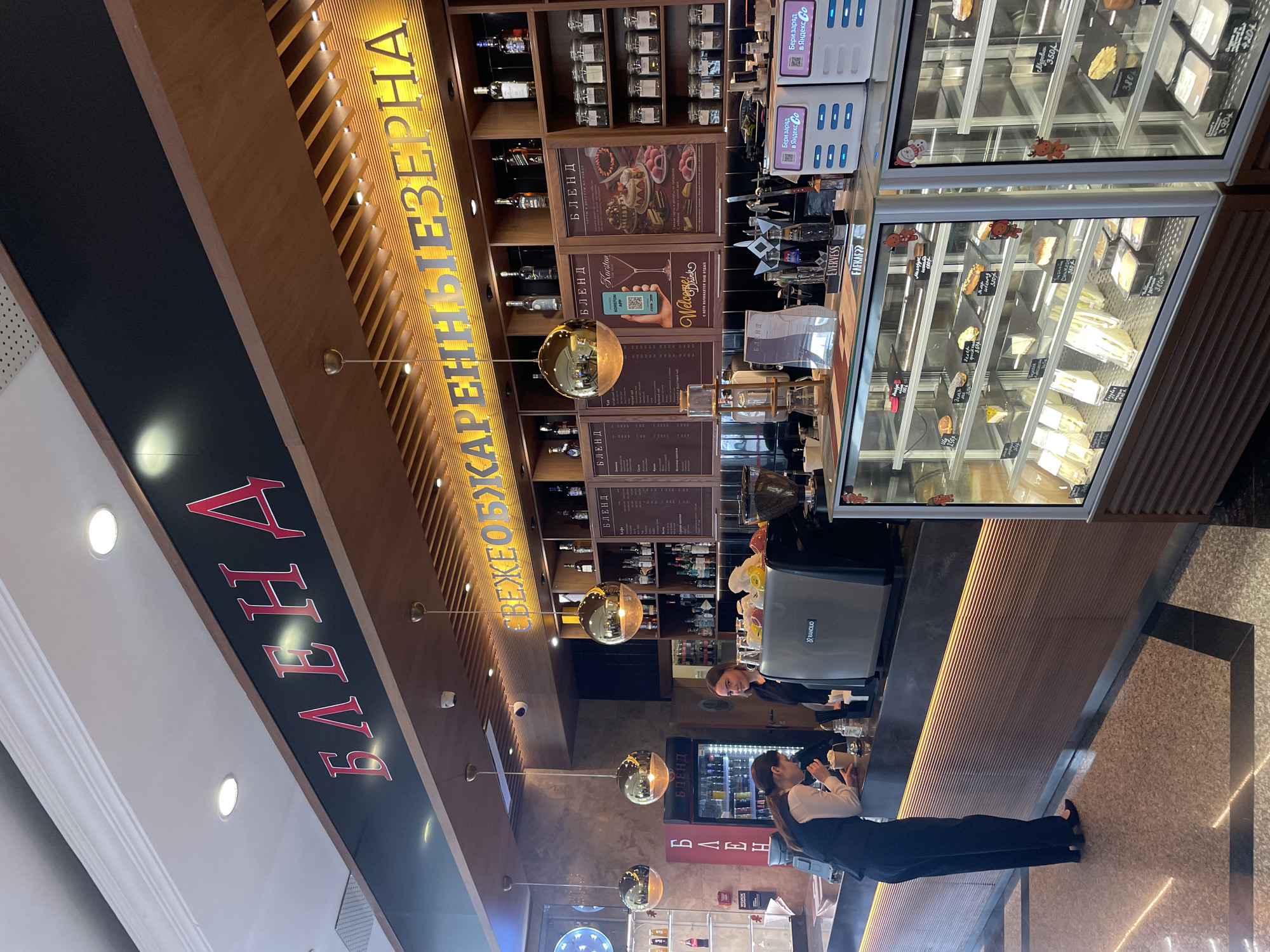 The image depicts the interior of a cafe named "БЛЕНД" (BLEND) located in Kazan, Russia. The cafe features a modern design with warm wood accents and ambient lighting. In the foreground, a glass display case showcases various pastries and sandwiches, with price tags indicating their cost in Russian Rubles. Two female employees are visible behind the counter. One employee, wearing a dark vest over a white shirt, is taking an order from a customer who is leaning on the counter and holding a cup. The other employee, also in dark attire, is standing behind the counter and looking towards the camera. The cafe's signage, with the name "БЛЕНД" in prominent red lettering, is visible on the wall above the counter. Behind the employees, shelves are stocked with bottles of alcohol and jars, suggesting it may also serve alcoholic beverages. The overall environment appears clean and well-lit, with a focus on displaying food and creating a welcoming atmosphere for customers.