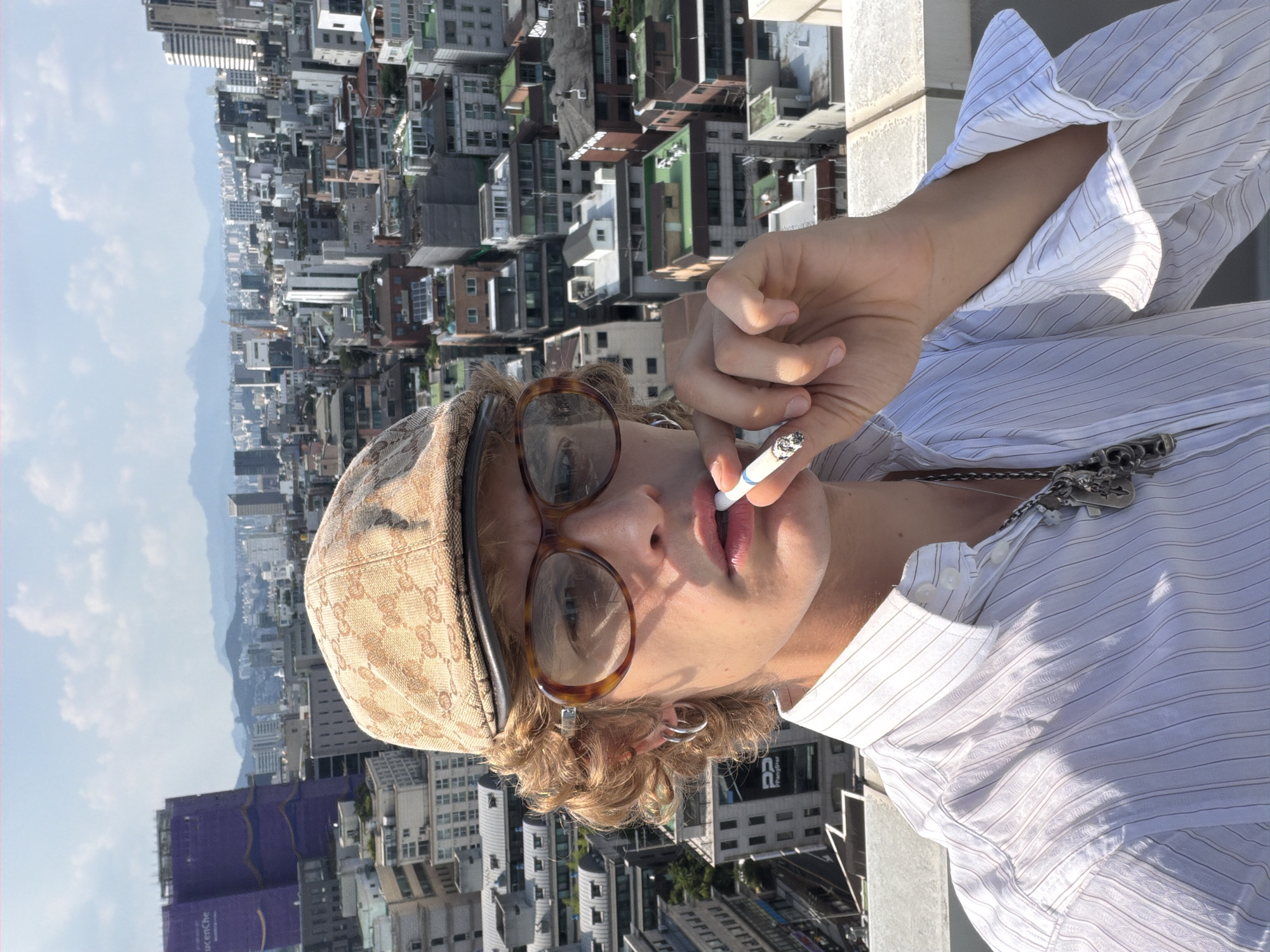 A young person is taking a selfie on a rooftop in Seoul, South Korea, with the cityscape in the background. They are wearing a light-colored, pinstriped button-up shirt and a tan flat cap with a repeating pattern. They have curly blonde hair, large tortoiseshell-framed glasses, and a silver hoop earring in their left ear. They are holding a lit cigarette to their lips with their right hand and are wearing a necklace with several silver charms. The sky is blue with some scattered clouds. The buildings in the background are a mix of modern and older architecture, with a large purple building visible on the left side of the image.