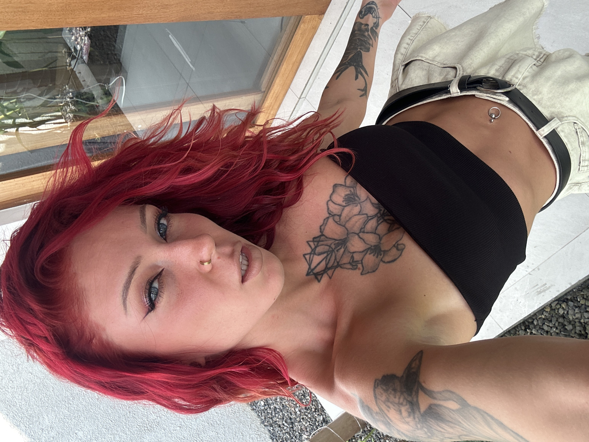 The image presents a woman with bright red hair, fair skin, and blue eyes, who appears to be positioned outdoors. She is wearing a black cropped top and light-colored, loose-fitting pants secured with a black belt. A small ring is visible in her nose, and she has a navel piercing. Several tattoos are prominently displayed on her chest and arms.
The background consists of what appears to be a modern building with large windows and landscaping featuring small stones. The location is suggested to be North Kuta, Indonesia, though this is not definitively verifiable from the image alone.
The woman is looking directly at the camera.