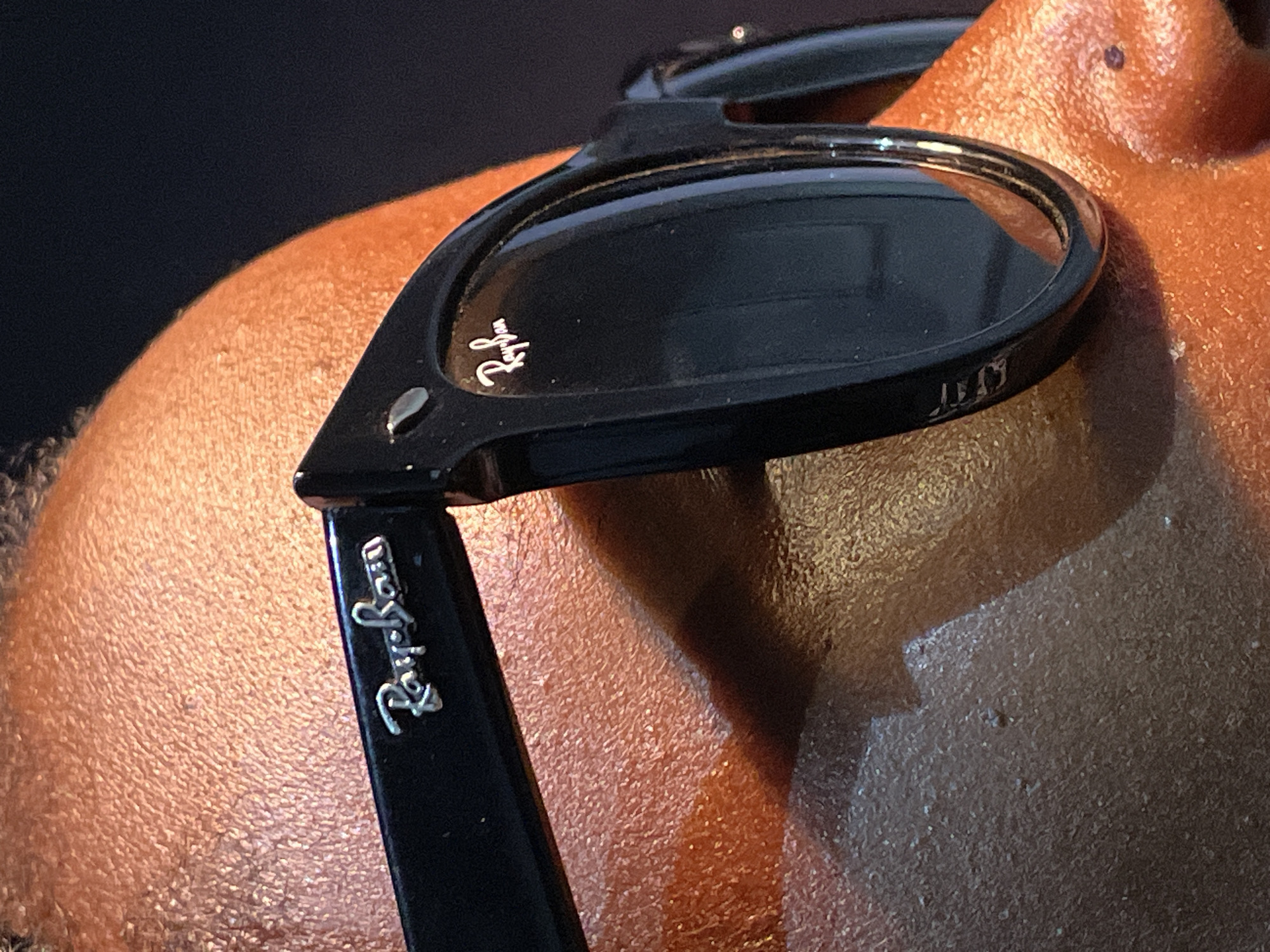 A close-up image focuses on a pair of black Ray-Ban sunglasses resting on a person's brown skin, likely the top of their head or shoulder. The "Ray-Ban" logo is prominently displayed on the side arm and faintly visible on the lens. The background is dark and out of focus. The image contains no discernible information about the city of Barakin Akawo, Nigeria.