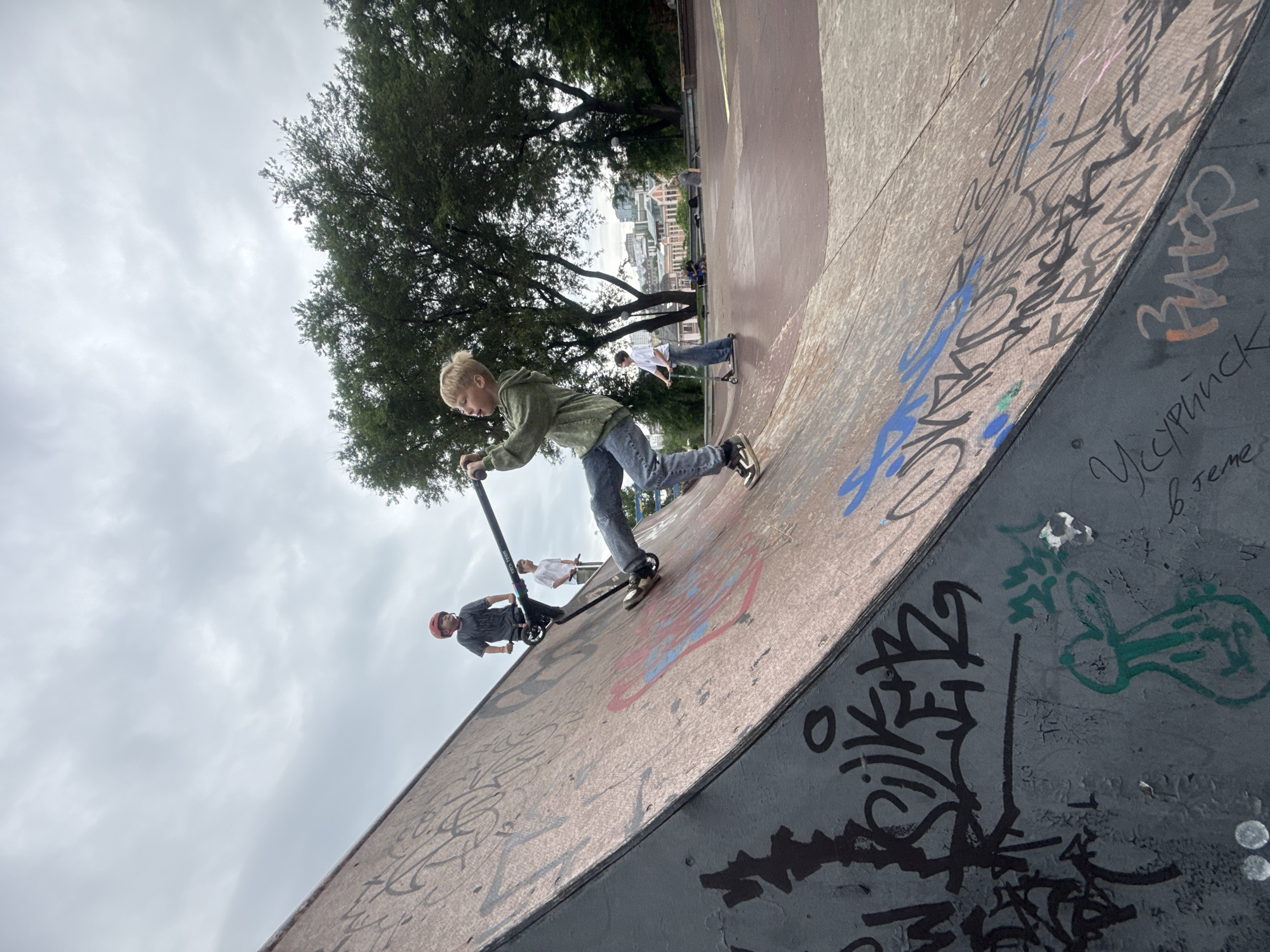A boy is skateboarding on a ramp with graffiti in Vladivostok, Russia. The boy is wearing a green hoodie and blue jeans, and he is holding a scooter. He is on a ramp in a skate park. There are other people in the background, also skateboarding or watching. The sky is cloudy, and there are trees in the background. The ramp is made of wood and concrete, and it is covered in graffiti.