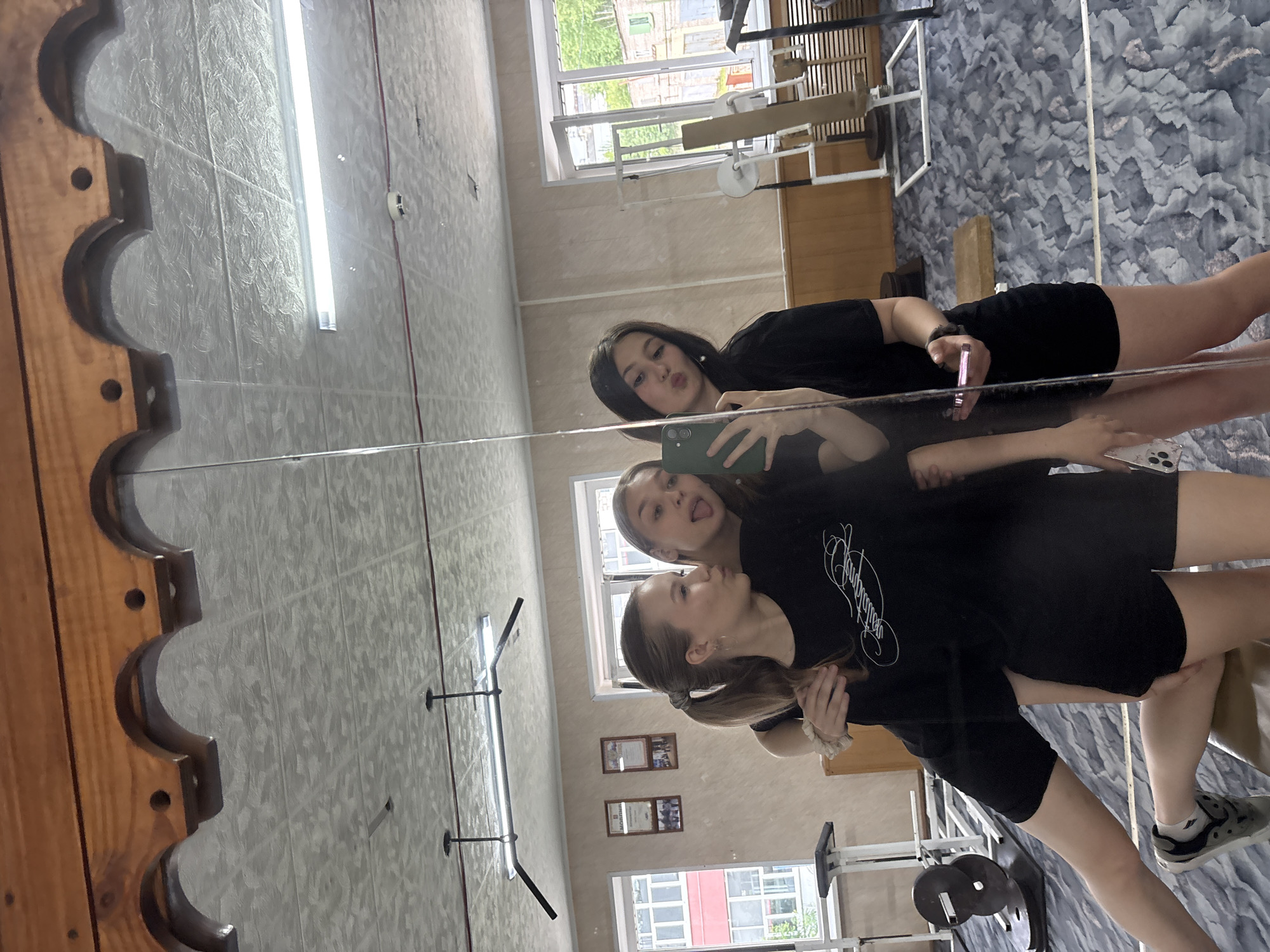 Three young women are taking a mirror selfie in what appears to be a gymnasium. The woman on the left is leaning forward, with her arm around the woman in the middle. The woman in the middle is sticking her tongue out, and the woman on the right is puckering her lips. They are all wearing black athletic clothing. In the background, there are windows, gym equipment, and a bulletin board. The floor is carpeted with a blue and gray abstract pattern. The ceiling has a textured tile pattern and fluorescent lighting. The overall impression is of a casual and playful moment.