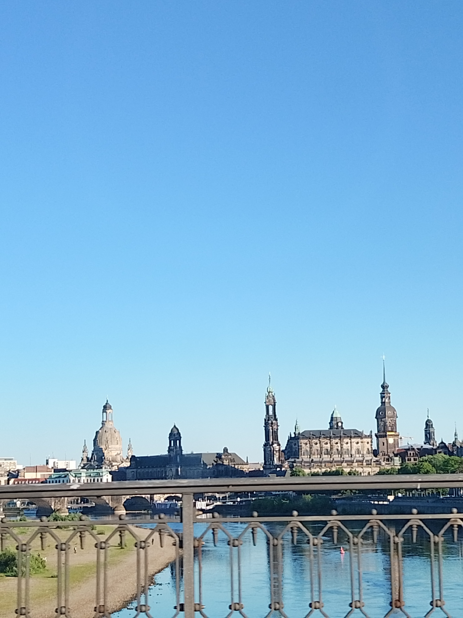 The image depicts a skyline of Dresden, Germany, viewed from a bridge over the Elbe river on a clear day. The skyline includes prominent buildings like the Frauenkirche and Dresden Cathedral. The water reflects the buildings and sky. A metal railing of the bridge is in the foreground.