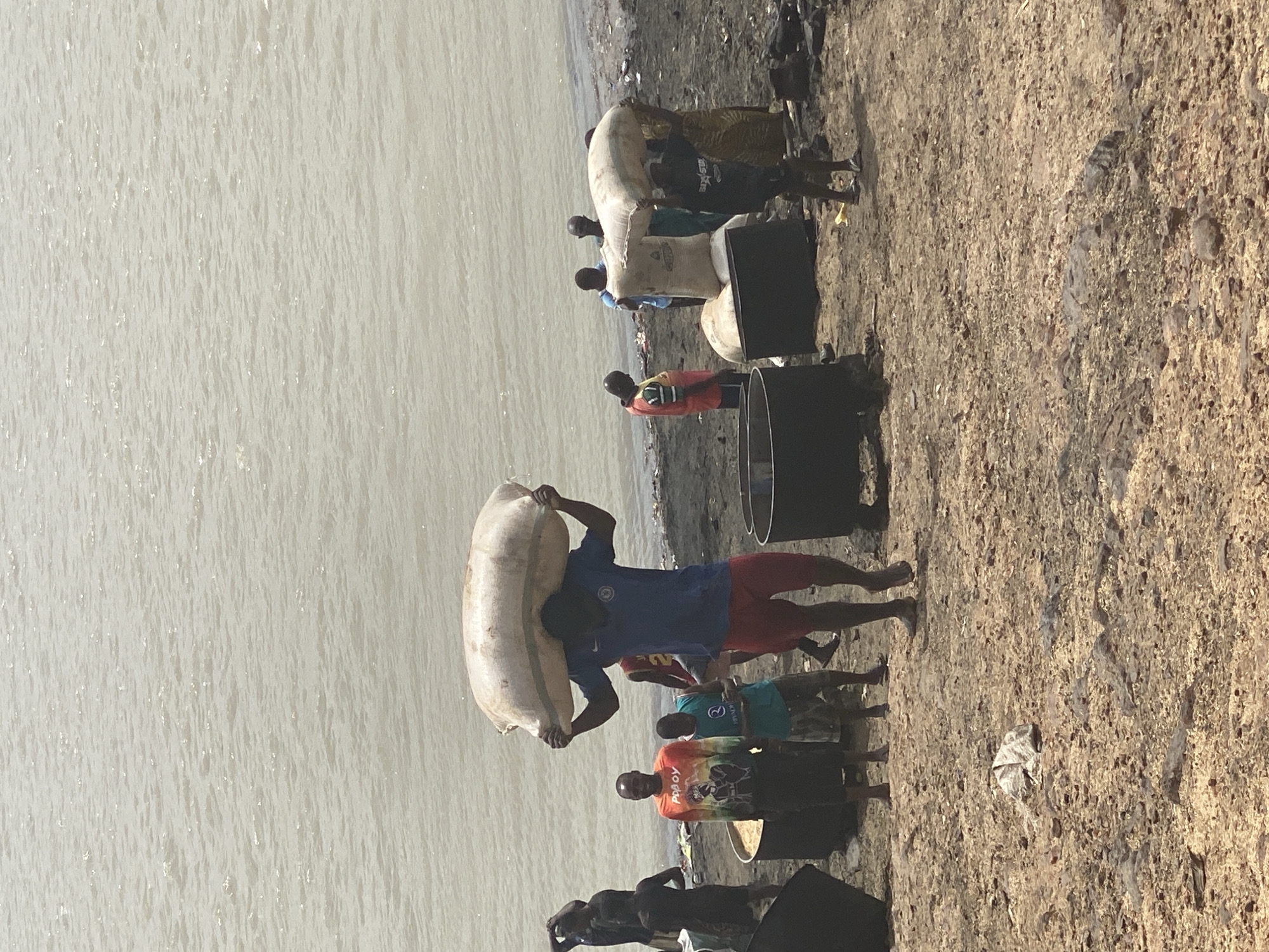The image shows a group of people working near a body of water in Lokoja, Nigeria. One man is carrying a large sack over his shoulder. Other individuals are standing near large metal containers and sacks. The ground is a mix of sand and debris. The body of water is calm and gray.